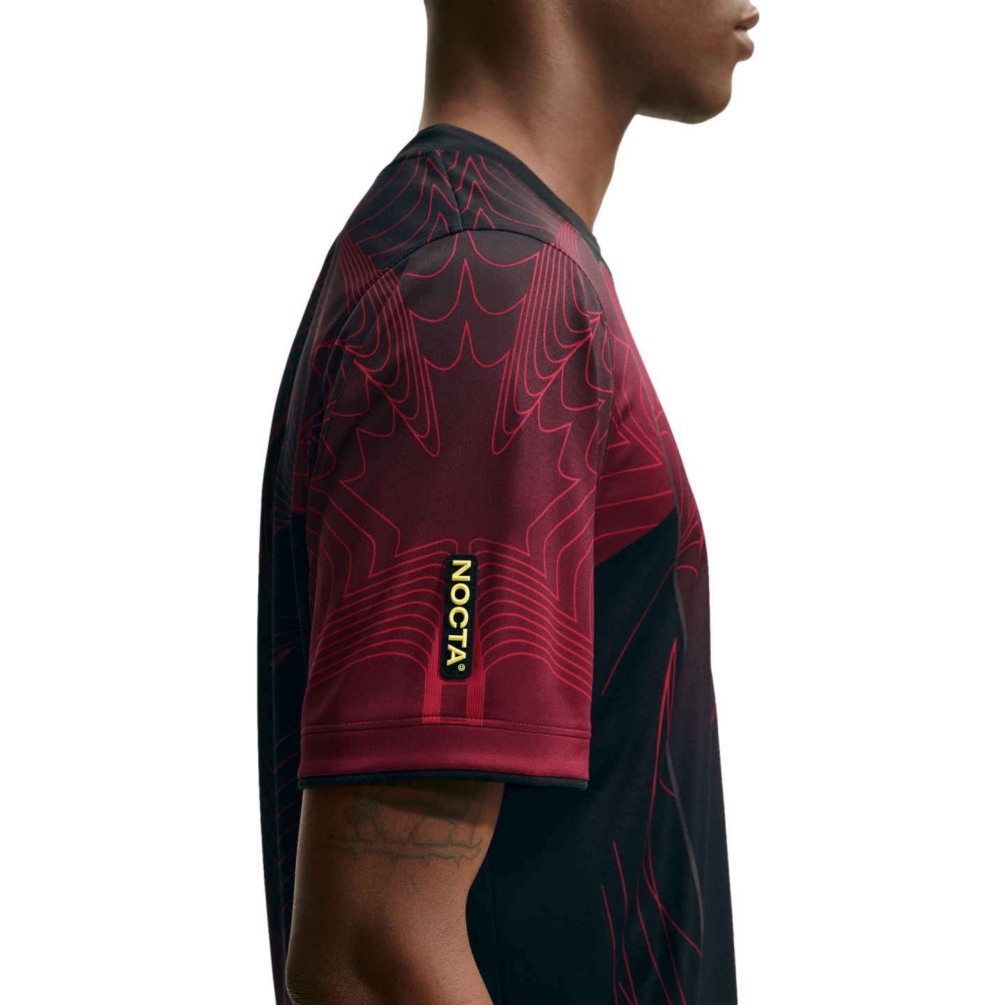 Side profile of a person wearing a maroon and black jersey with graphic line patterns and a yellow "NOCTA" label on the sleeve.
