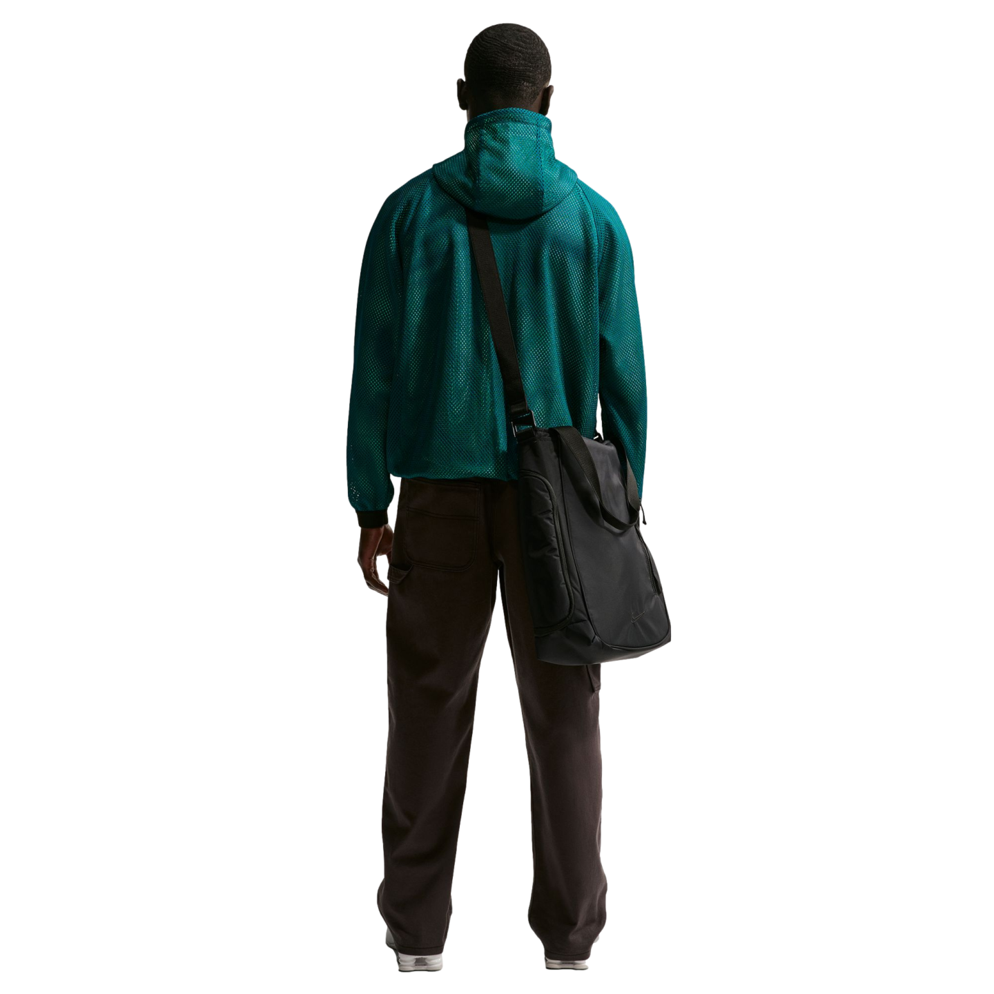 Rear view of a man standing against a solid black background, wearing a teal green mesh hooded jacket, dark brown trousers, and white sneakers. A large black messenger bag is slung over his right shoulder.