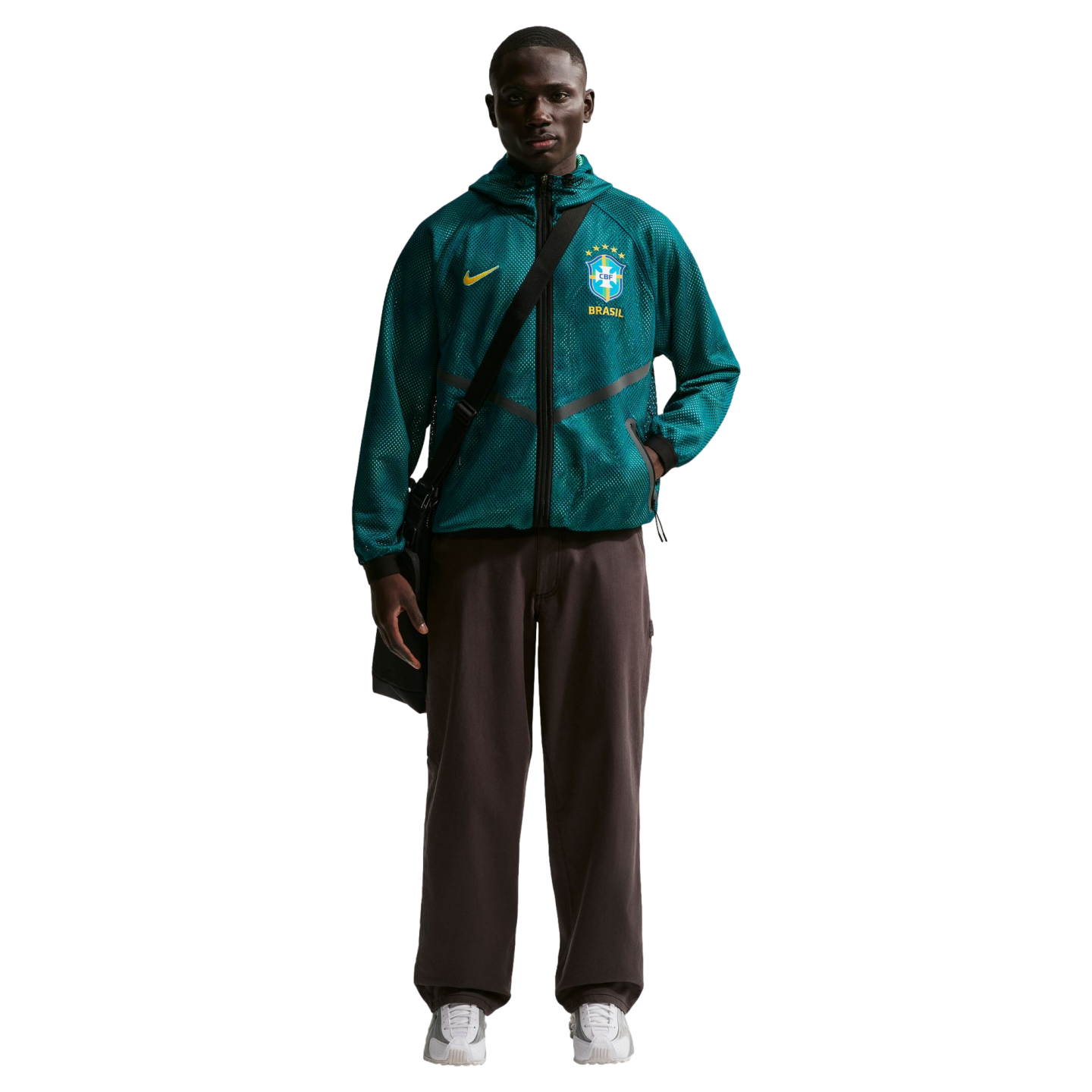 A young Black man stands against a black background wearing a teal mesh Nike zip-up hoodie featuring the Brazil national football team crest, dark brown wide-leg trousers, and white sneakers. He has a black messenger bag slung across his chest.