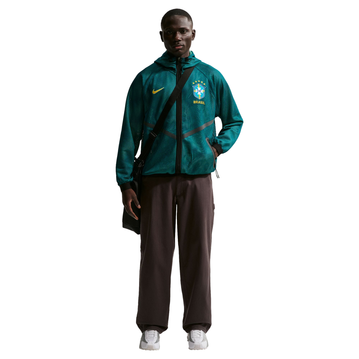 A young Black man stands against a black background wearing a teal mesh Nike zip-up hoodie featuring the Brazil national football team crest, dark brown wide-leg trousers, and white sneakers. He has a black messenger bag slung across his chest.