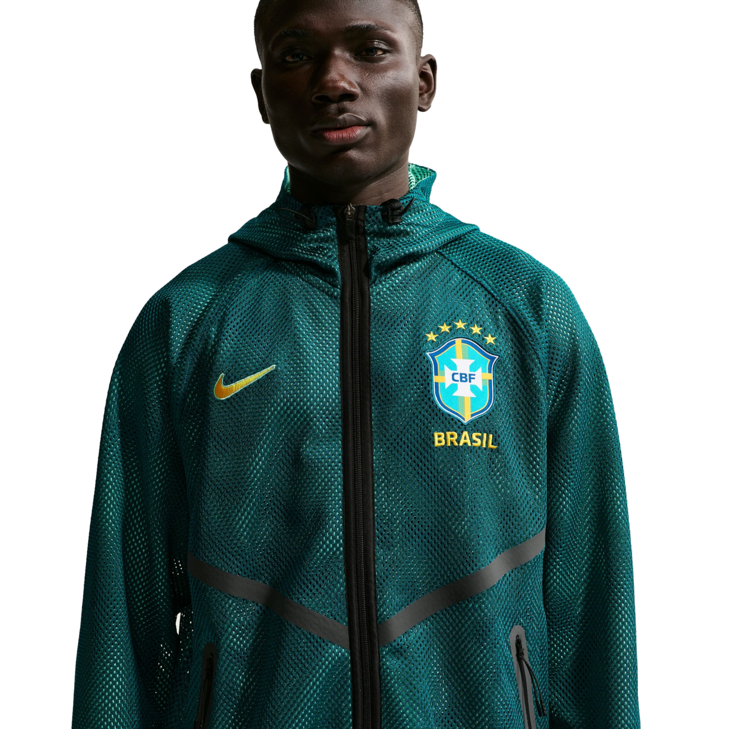 A waist-up portrait of a young Black man wearing a dark teal mesh Nike zip-up hoodie. The hoodie features a yellow Nike swoosh on one side and the Brazil national football team crest with "BRASIL" printed below it on the other. He is looking directly at t