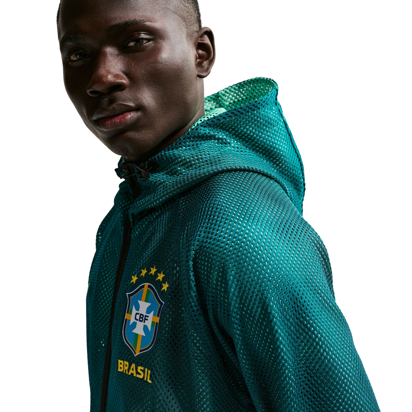 A close-up shot of a man with dark skin looking towards the camera against a solid black background. He is wearing a teal, mesh, zip-up hoodie with the Brazilian football confederation (CBF) crest on the chest, featuring five yellow stars above it and the