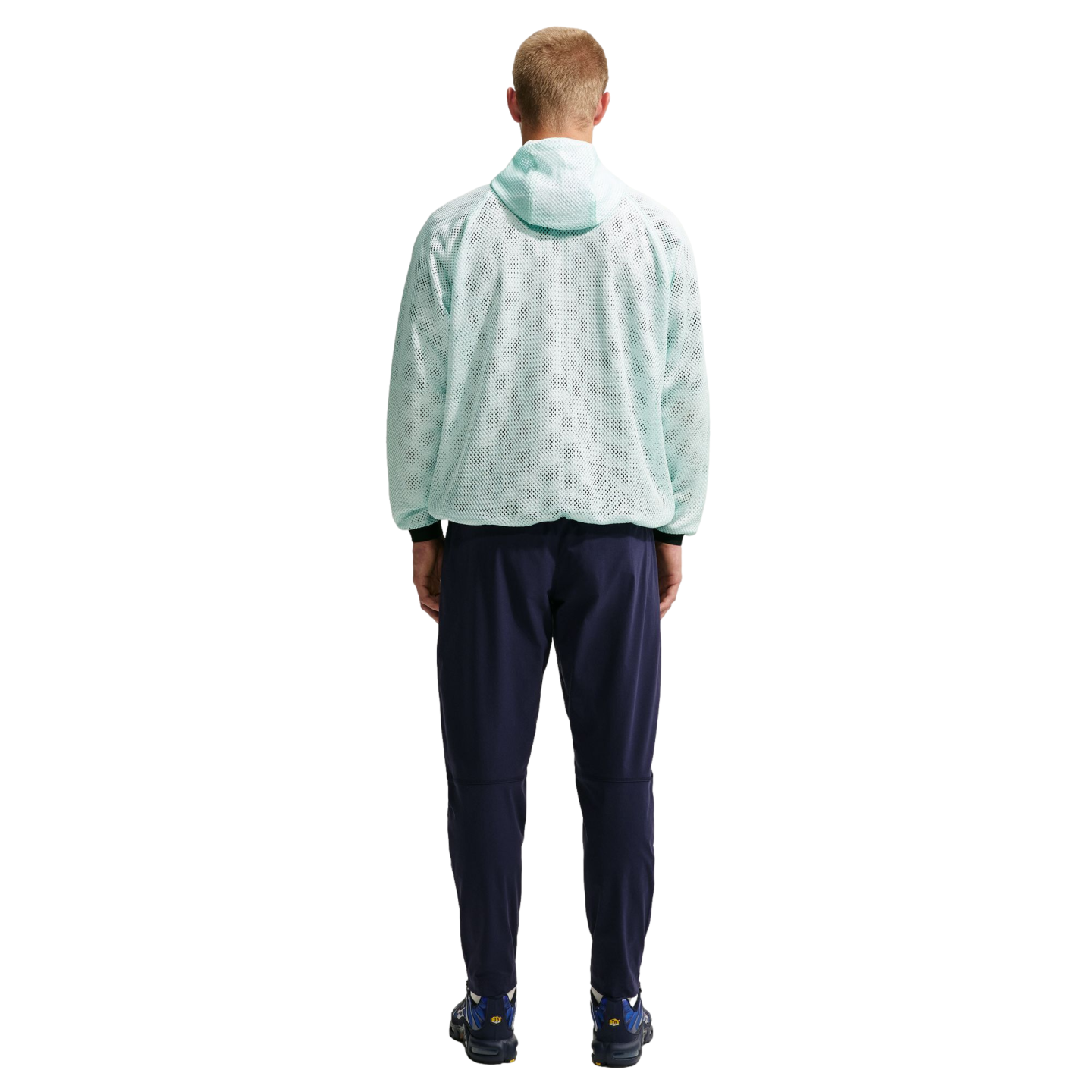 A man stands with his back to the camera, wearing a light mint green mesh hoodie, navy blue pants, and blue sneakers against a black background.