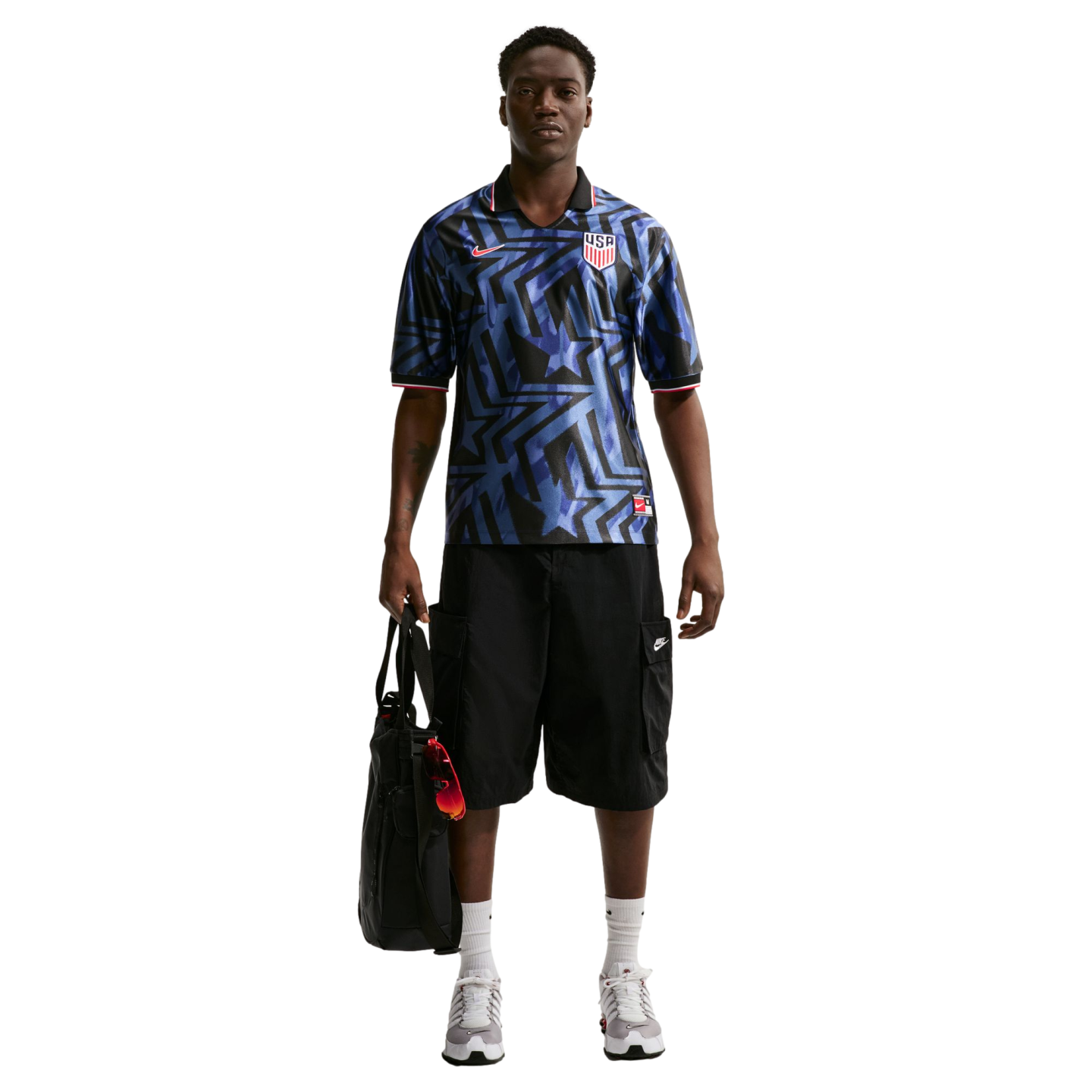 A Black man wearing a blue patterned USA soccer jersey, black cargo shorts, and sneakers stands holding a black bag against a white background.