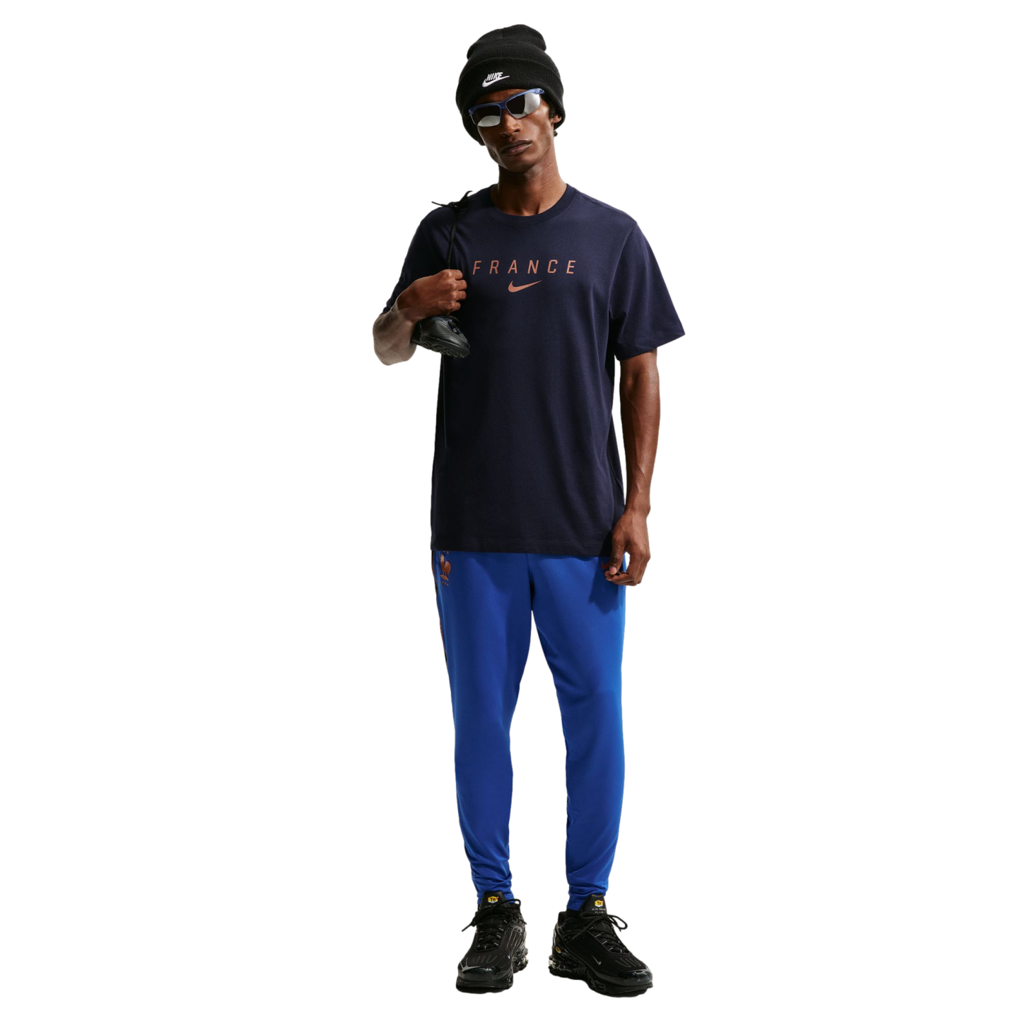A full-body photo of a man standing against a solid black background. He is wearing a dark navy blue Nike t-shirt with "FRANCE" printed on the chest, bright blue athletic pants, black sneakers, a black Nike beanie, and blue-tinted sunglasses. He is holdin