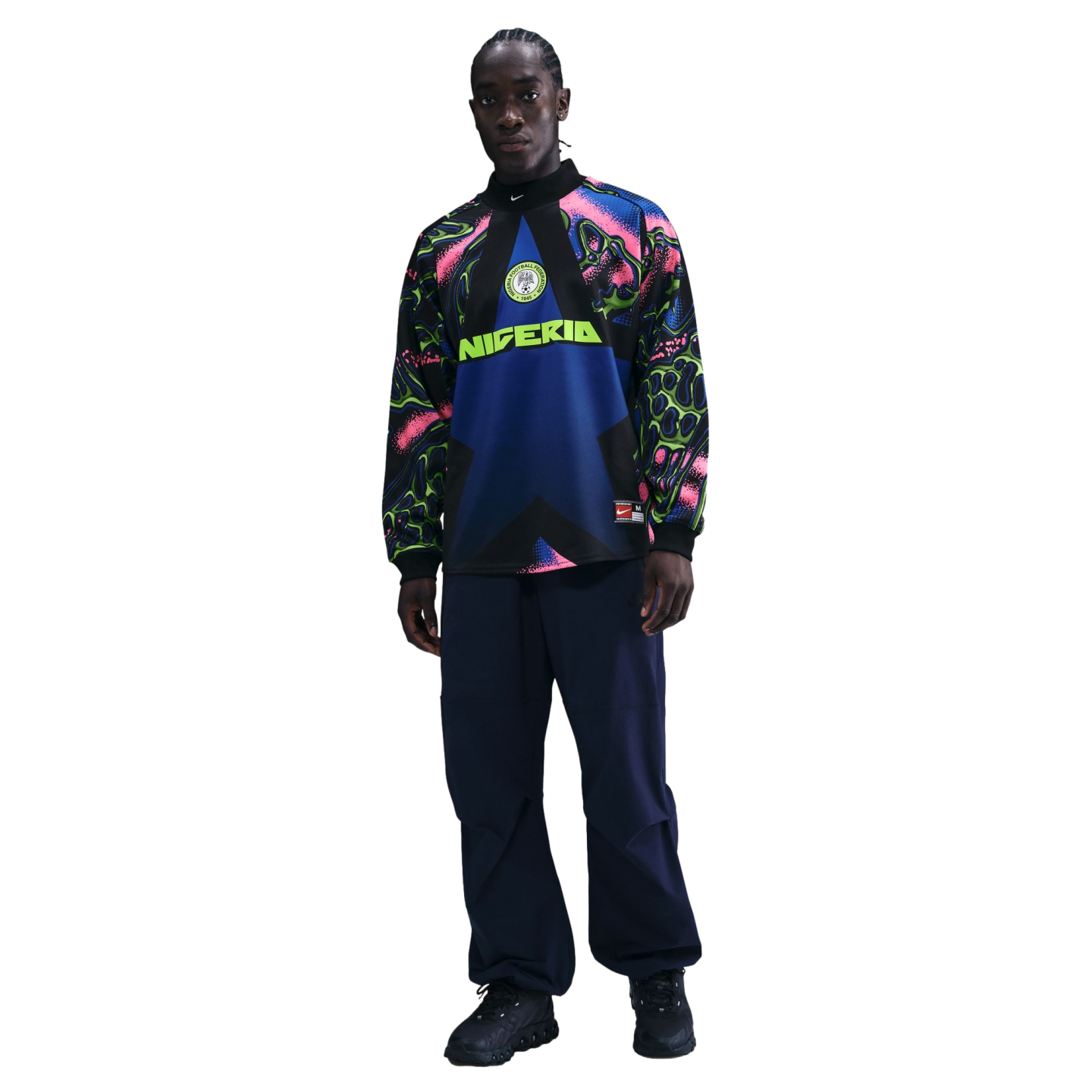 A man models a long-sleeve Nike Nigeria soccer top with a colorful abstract pattern on the sleeves and navy pants against a solid black background.