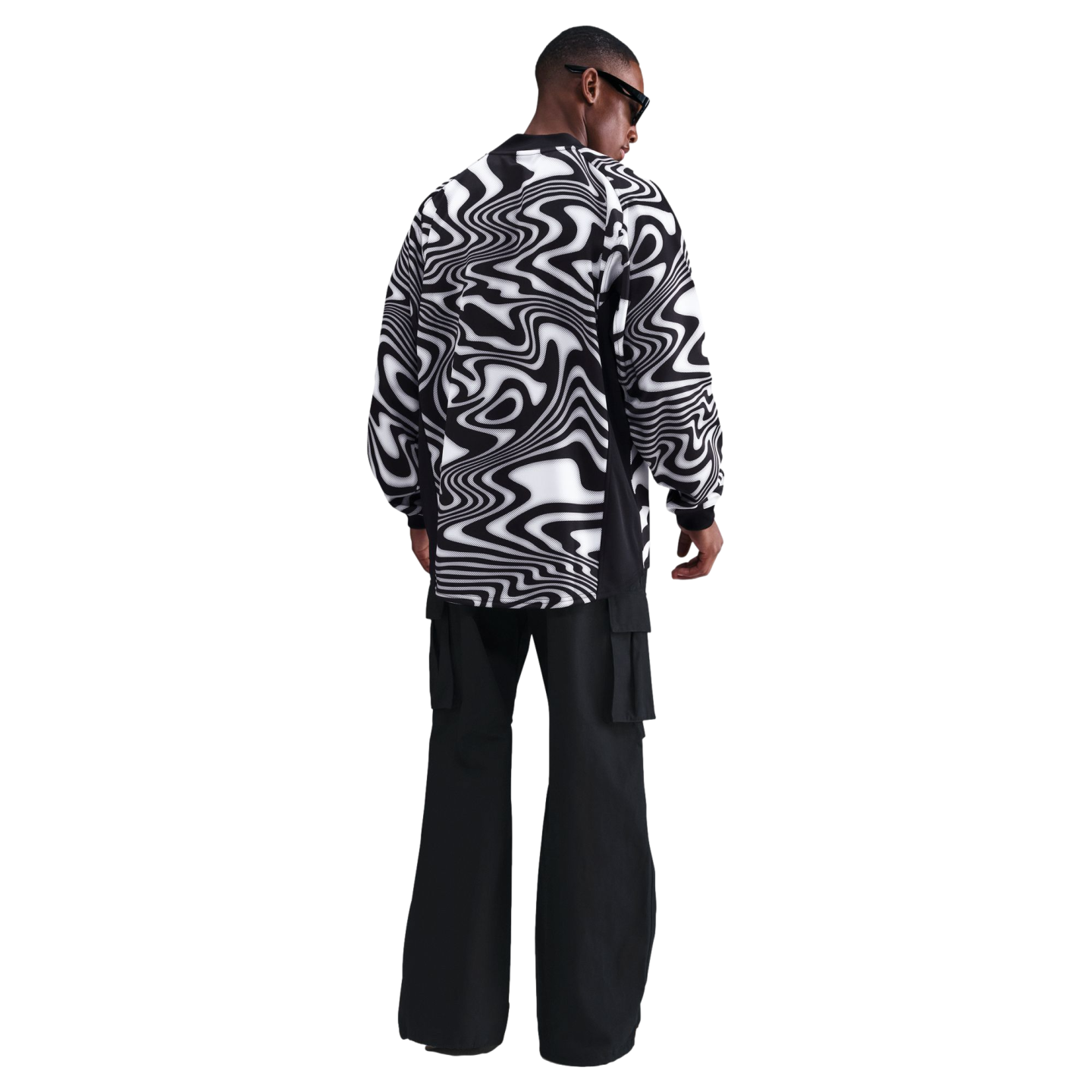 A man seen from behind wearing a black and white psychedelic-print long-sleeved shirt and black cargo pants against a black background.