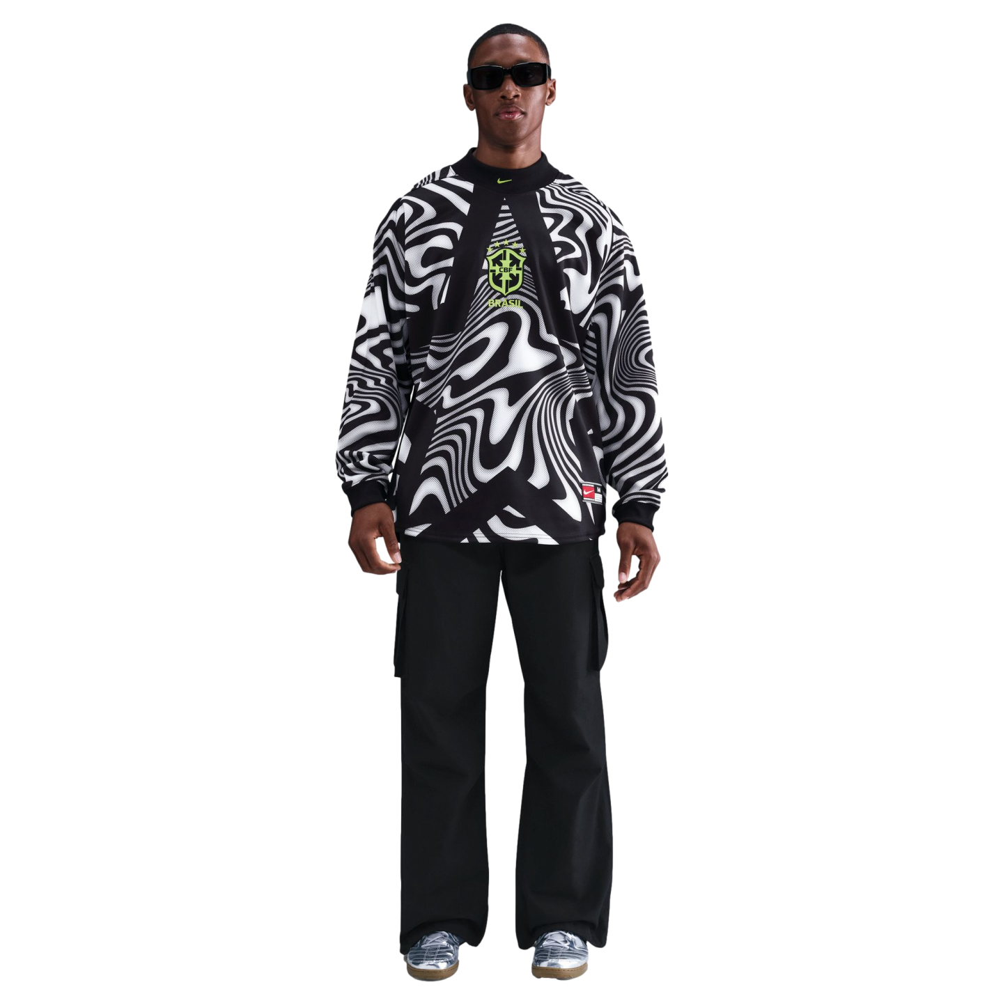 A man wearing sunglasses, a black and white wavy patterned Brazil top, black cargo pants, and silver sneakers against a black background.