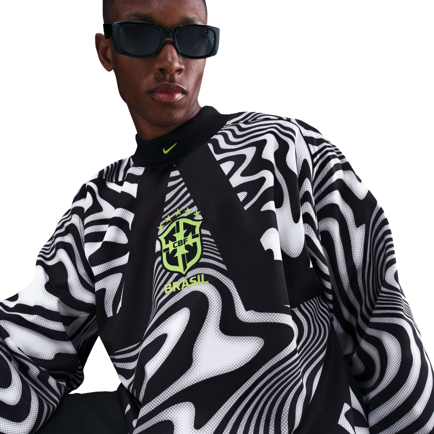 A person in black sunglasses wearing a high-neck long-sleeve shirt with a wavy black-and-white pattern and a lime green Brasil crest, against a black background.