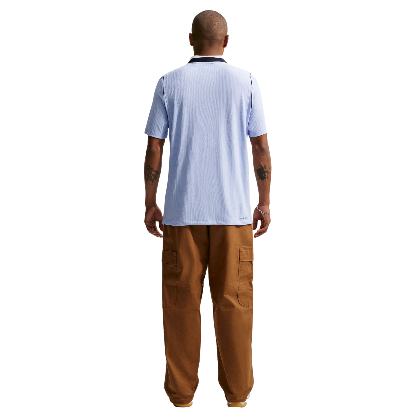 A man standing with his back to the camera against a solid black background, wearing a light blue ribbed short-sleeved shirt and baggy brown cargo pants. He has tattoos on both of his forearms.