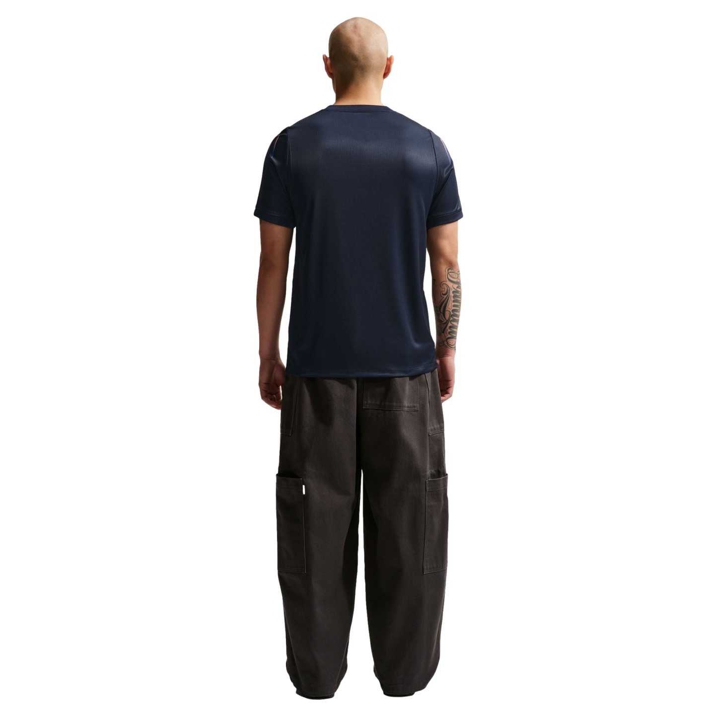 A rear view of a bald man wearing a navy blue t-shirt and oversized dark cargo pants, standing against a solid black background.