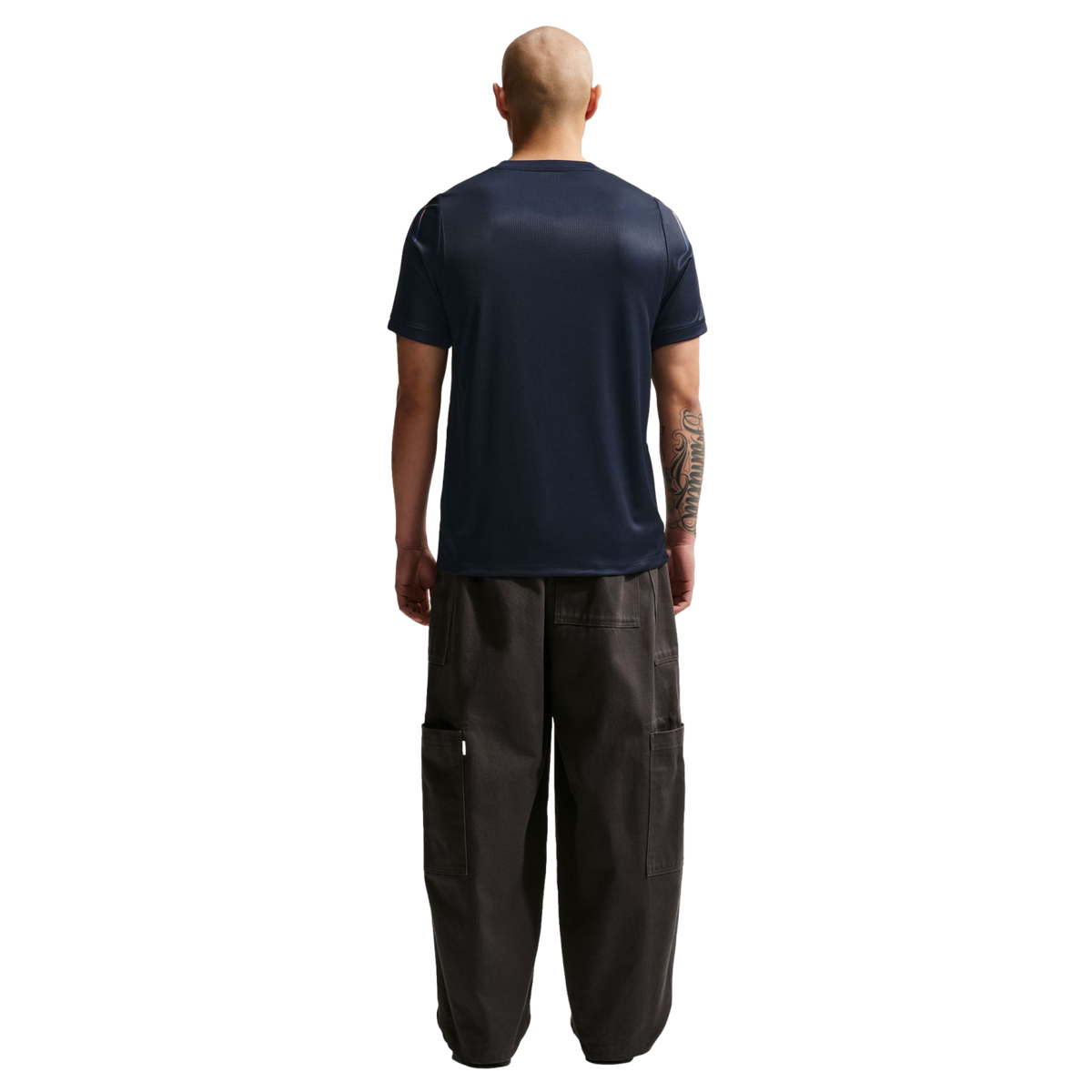 A rear view of a bald man wearing a navy blue t-shirt and oversized dark cargo pants, standing against a solid black background.