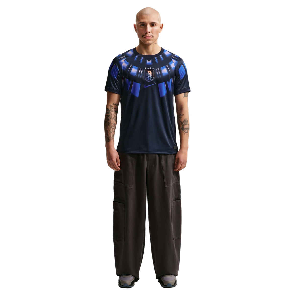 A full-body studio portrait of a bald man with arm tattoos standing against a solid black background. He is wearing a dark navy blue Nike t-shirt featuring a bright blue radial graphic around the collar and a center-chest Uruguay national team crest. His