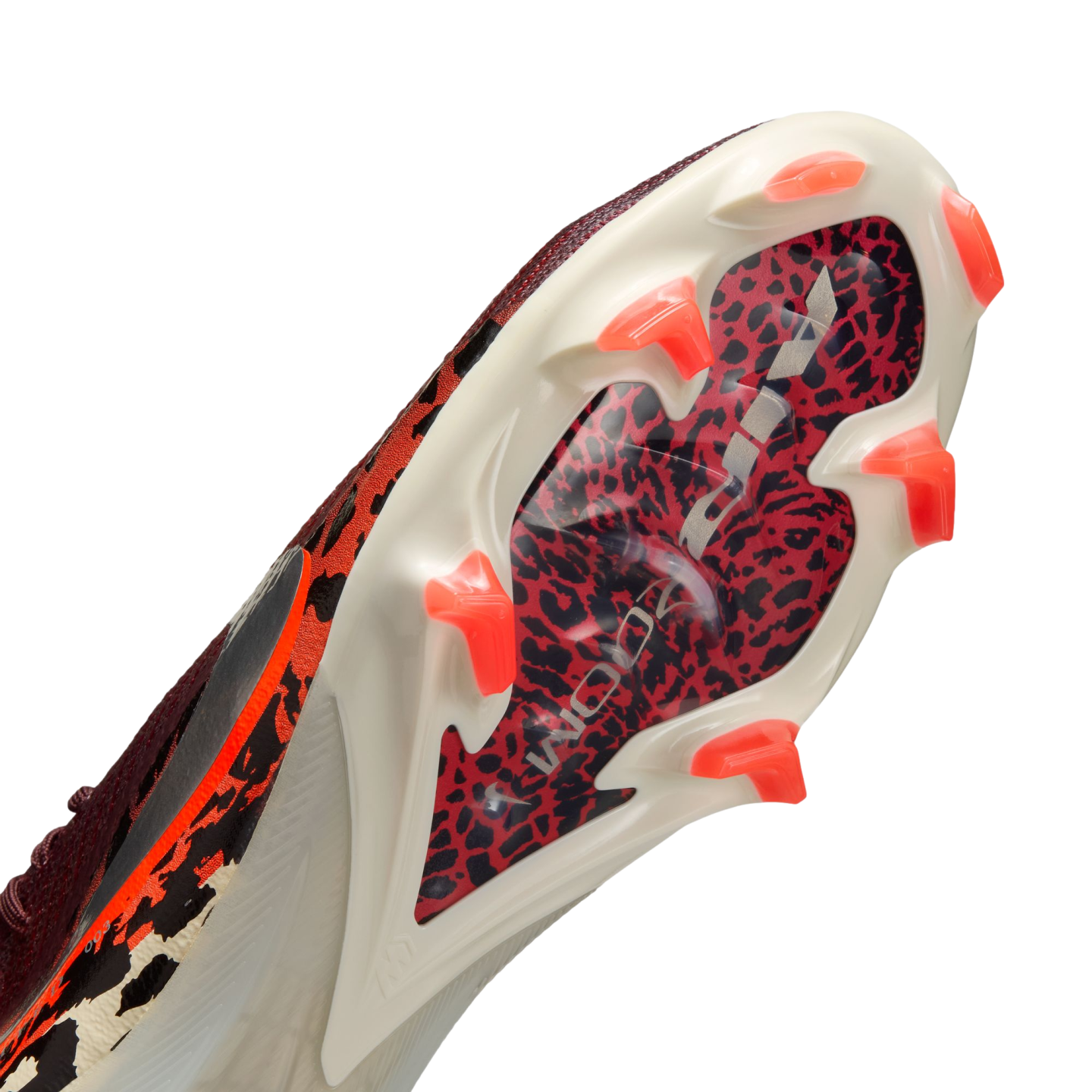 An angled, close-up view of the soleplate of a white soccer cleat, featuring a red and black leopard print pattern under a clear section with silver "AIR ZOOM" branding. The cleat has bright orange, translucent, chevron-shaped studs.