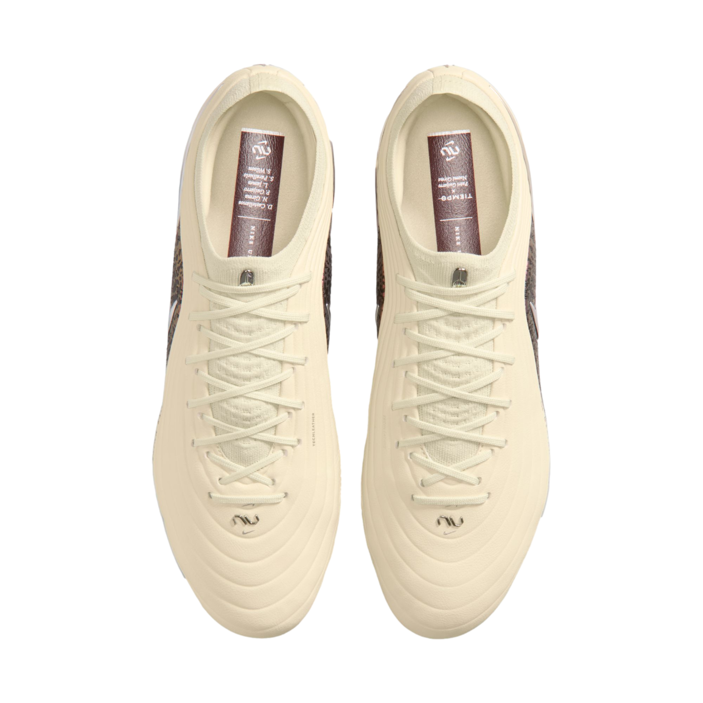 A top-down view of a pair of off-white Nike soccer cleats against a solid black background. The cleats feature cream-colored uppers and laces, with dark reddish-brown insoles containing white text and logos.