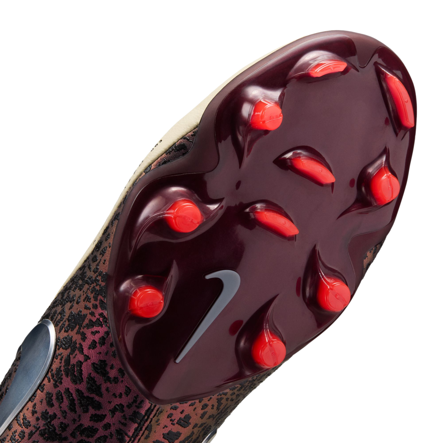 A close-up view of the glossy burgundy outsole of a Nike soccer cleat, featuring bright red studs and a grey swoosh logo. The textured upper of the shoe shows a dark, bronze-colored animal-print pattern.