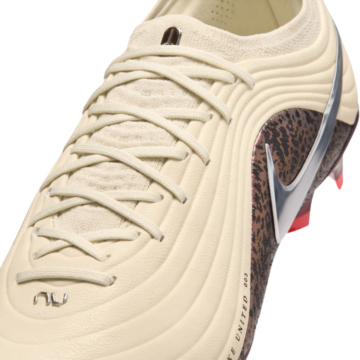 A close-up shot of a cream-colored Nike soccer cleat with a textured ribbed upper and matching laces. A metallic silver swoosh sits atop a dark brown mottled pattern on the side. The background is solid black.