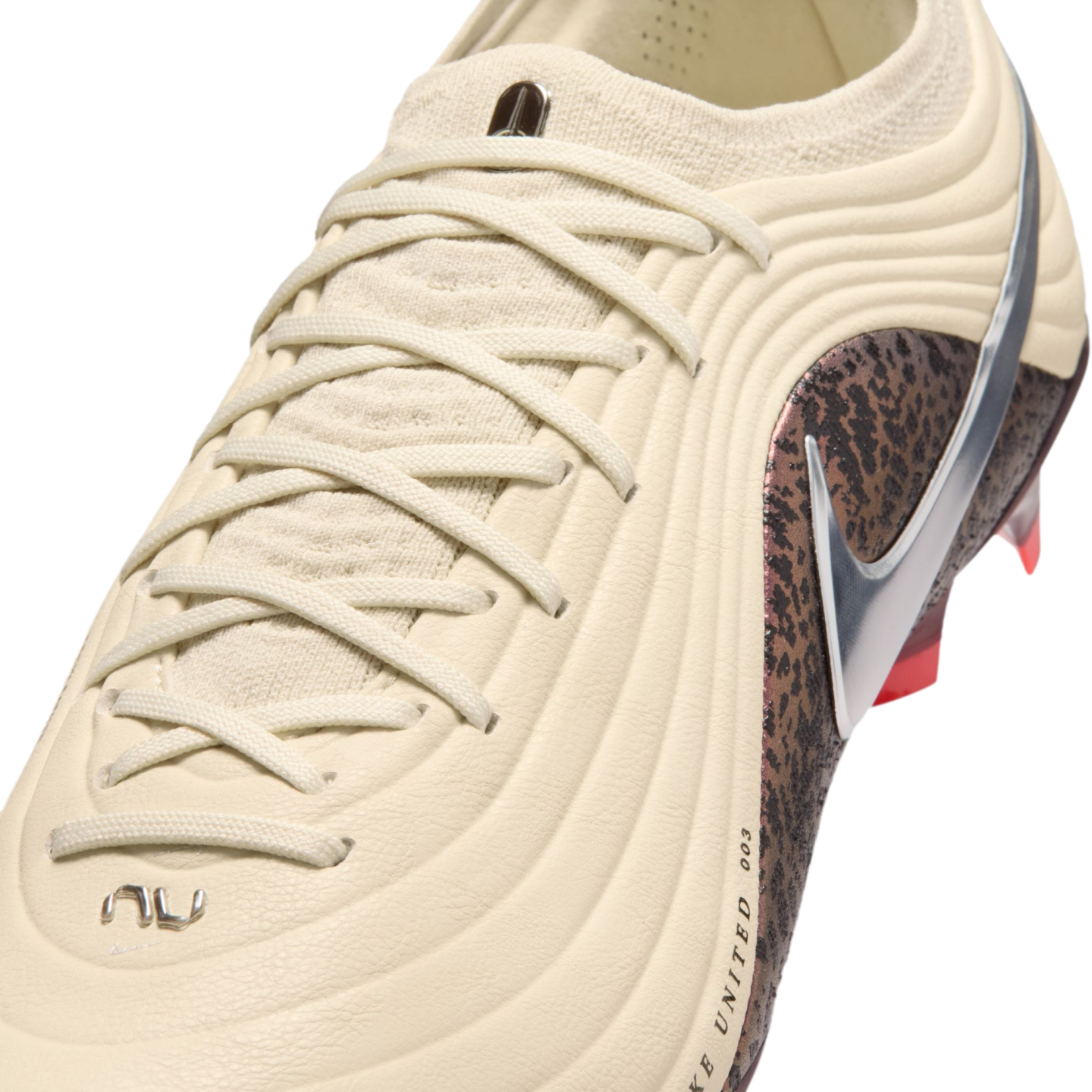 A close-up shot of a cream-colored Nike soccer cleat with a textured ribbed upper and matching laces. A metallic silver swoosh sits atop a dark brown mottled pattern on the side. The background is solid black.