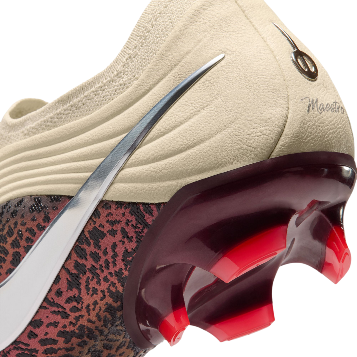 A close-up view of the heel and soleplate of a Nike soccer cleat against a black background. The upper features a cream-colored textured heel with a script "Maestro" inscription and a silver metallic Nike Swoosh. A glossy, deep-red translucent heel counte