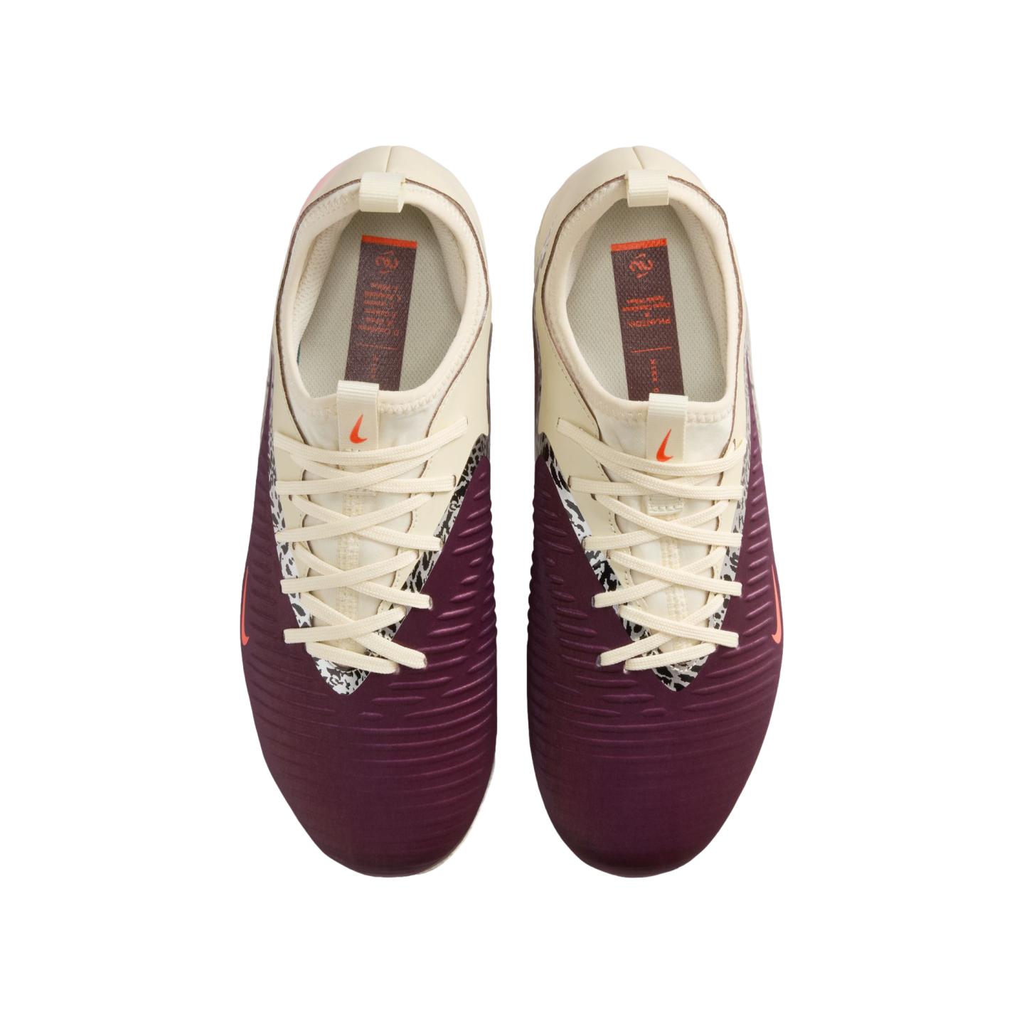 A top-down view of a pair of Nike football boots against a black background. The shoes feature a maroon lower section with horizontal ridges and a cream upper section. A black-and-white patterned trim lines the lace area, and small orange Nike swooshes ar