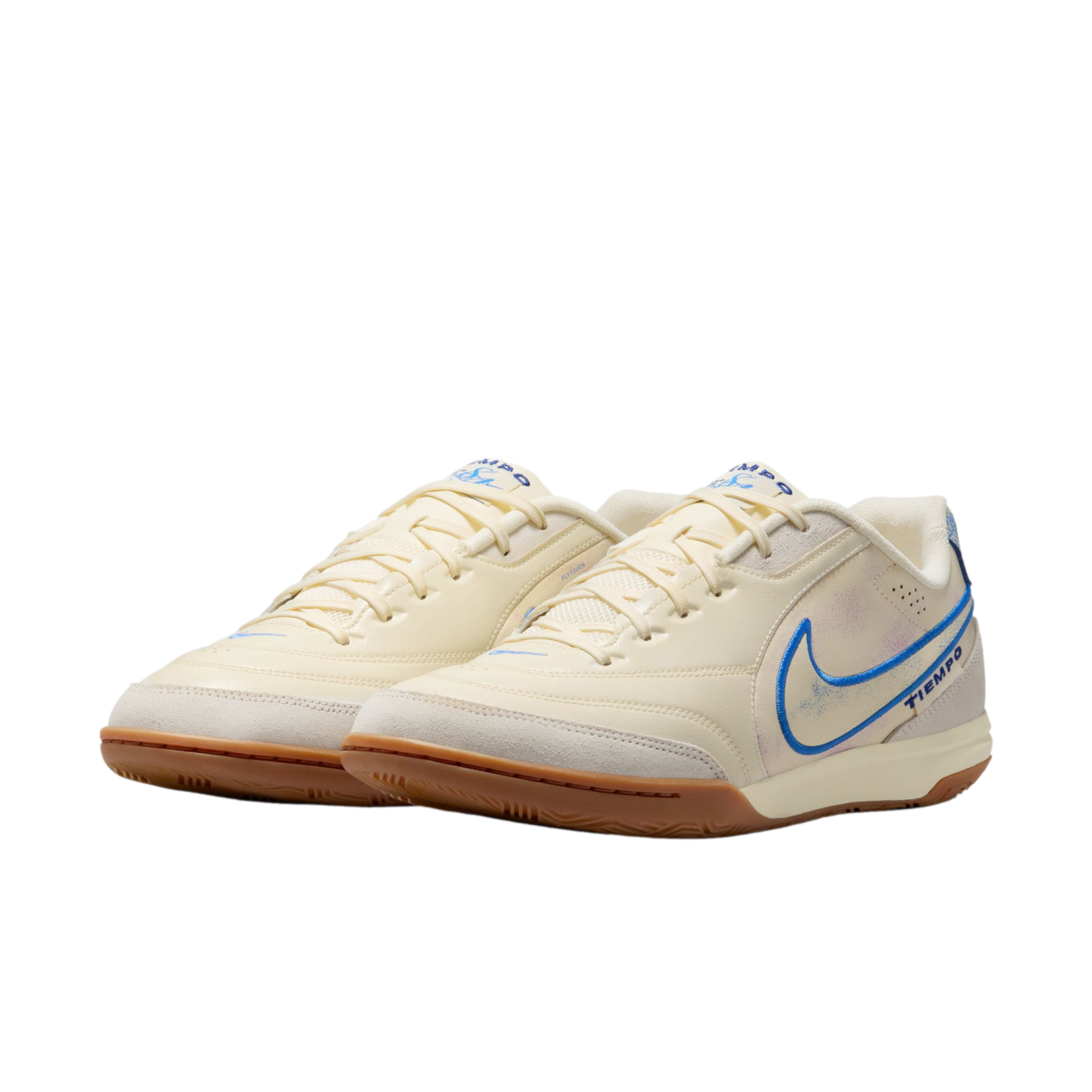 A pair of cream-colored Nike Tiempo indoor soccer shoes with blue accents and gum rubber soles, set against a black background.