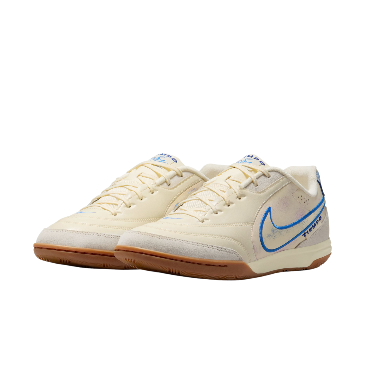 A pair of cream-colored Nike Tiempo indoor soccer shoes with blue accents and gum rubber soles, set against a black background.