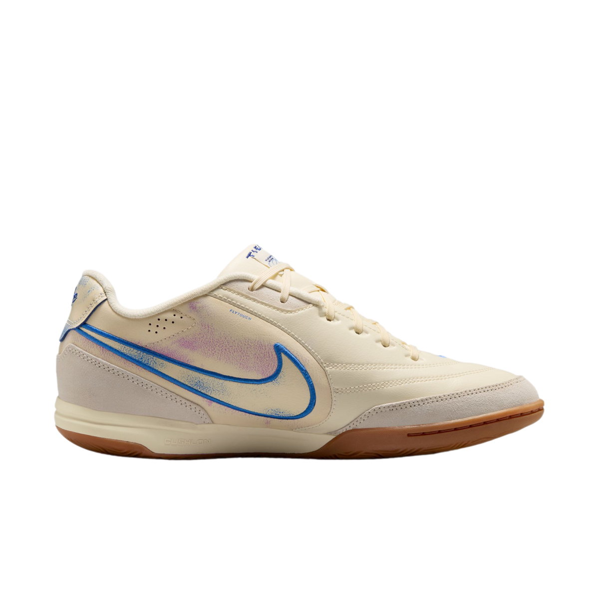 A side profile of a cream-colored Nike indoor soccer shoe against a black background, featuring a blue swoosh with purple accents and a brown gum sole.
