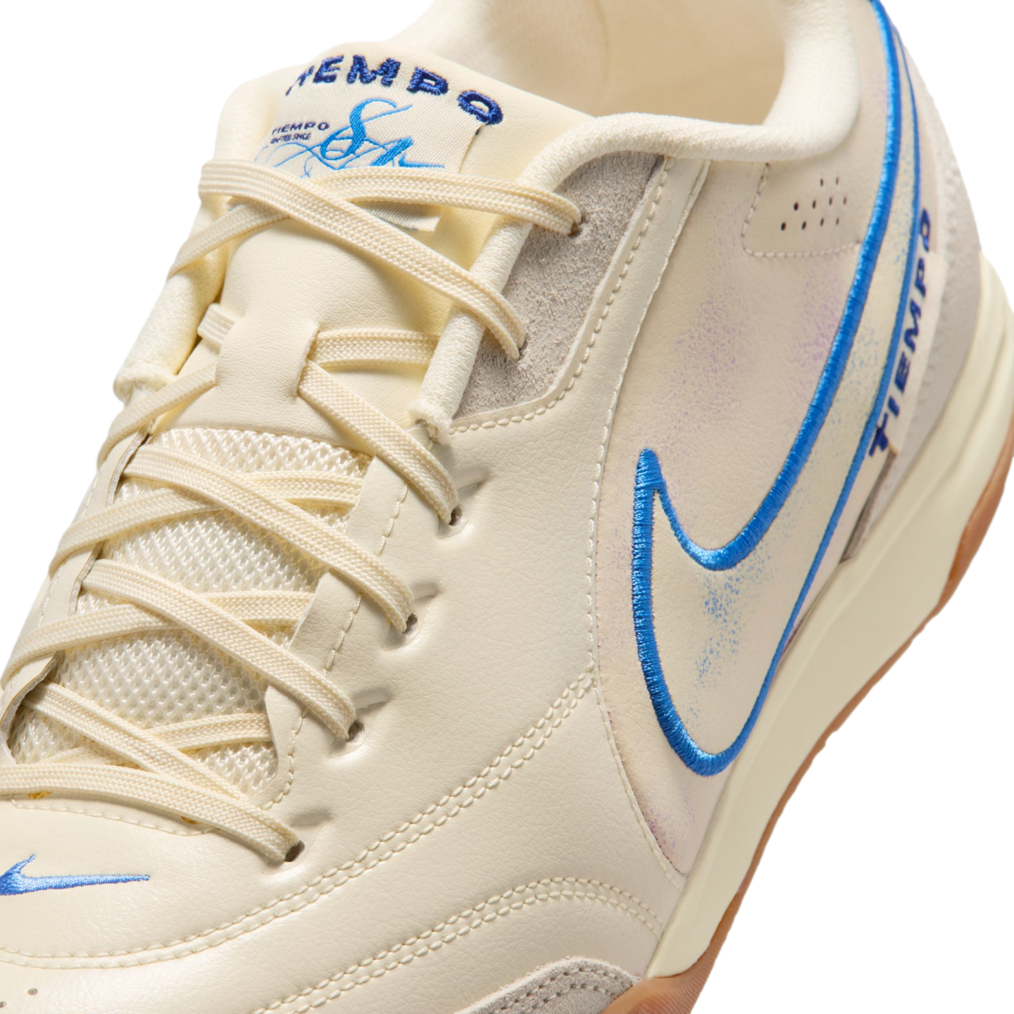 A close-up, angled shot of a cream-colored Nike Tiempo sneaker with blue embroidered accents, matching laces, and subtle iridescent details on the side.
