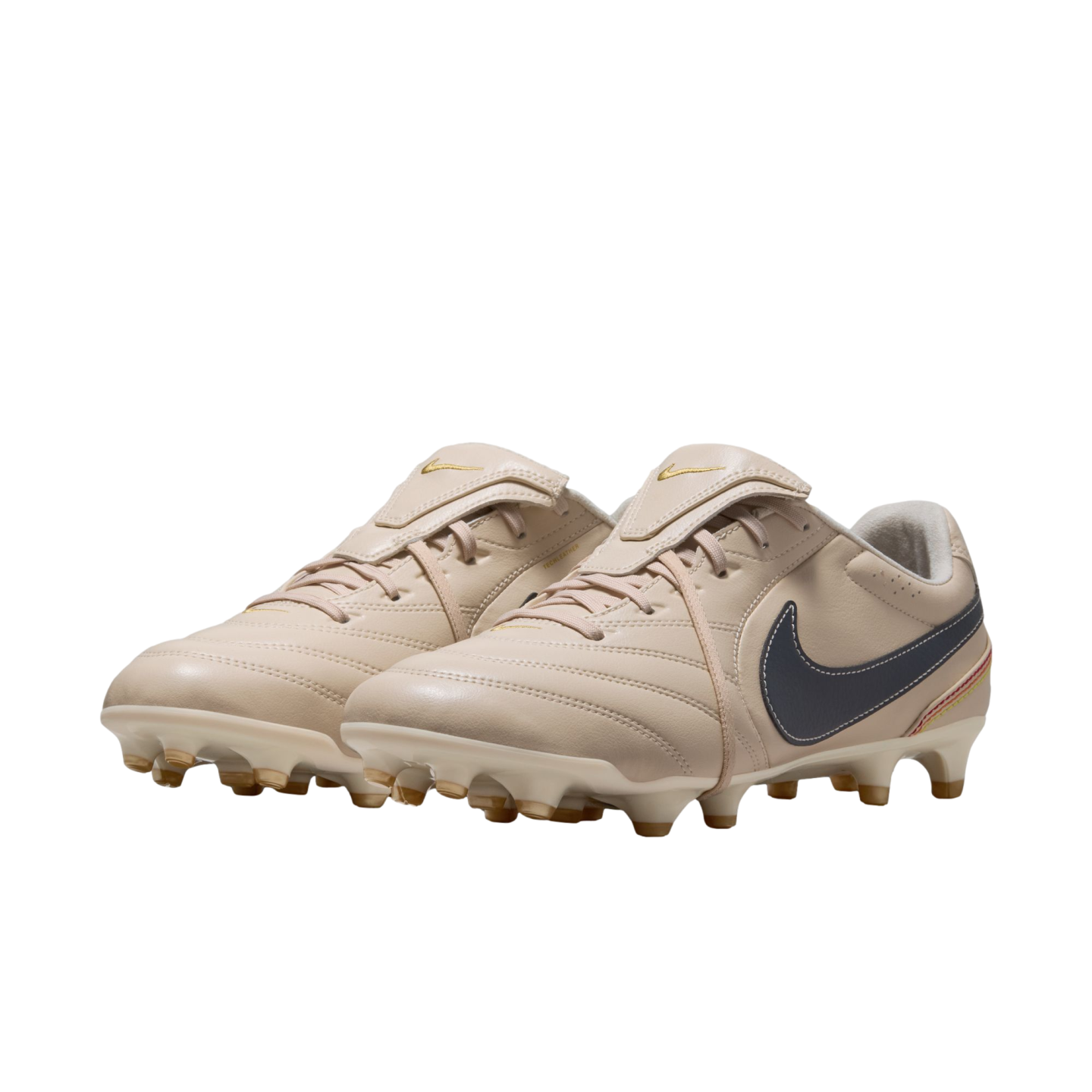 A pair of cream-colored Nike soccer cleats with a dark grey swoosh and gold accents, shown against a solid black background.