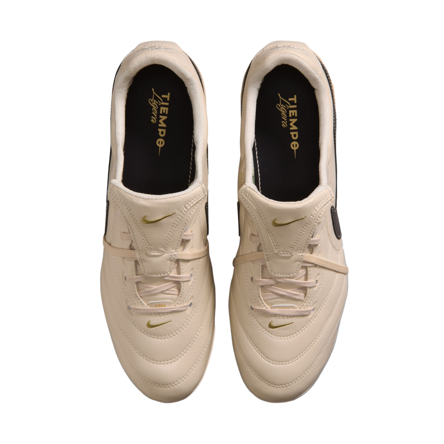 A top-down view of a pair of cream-colored Nike Tiempo Ligera soccer shoes with gold accents against a black background.