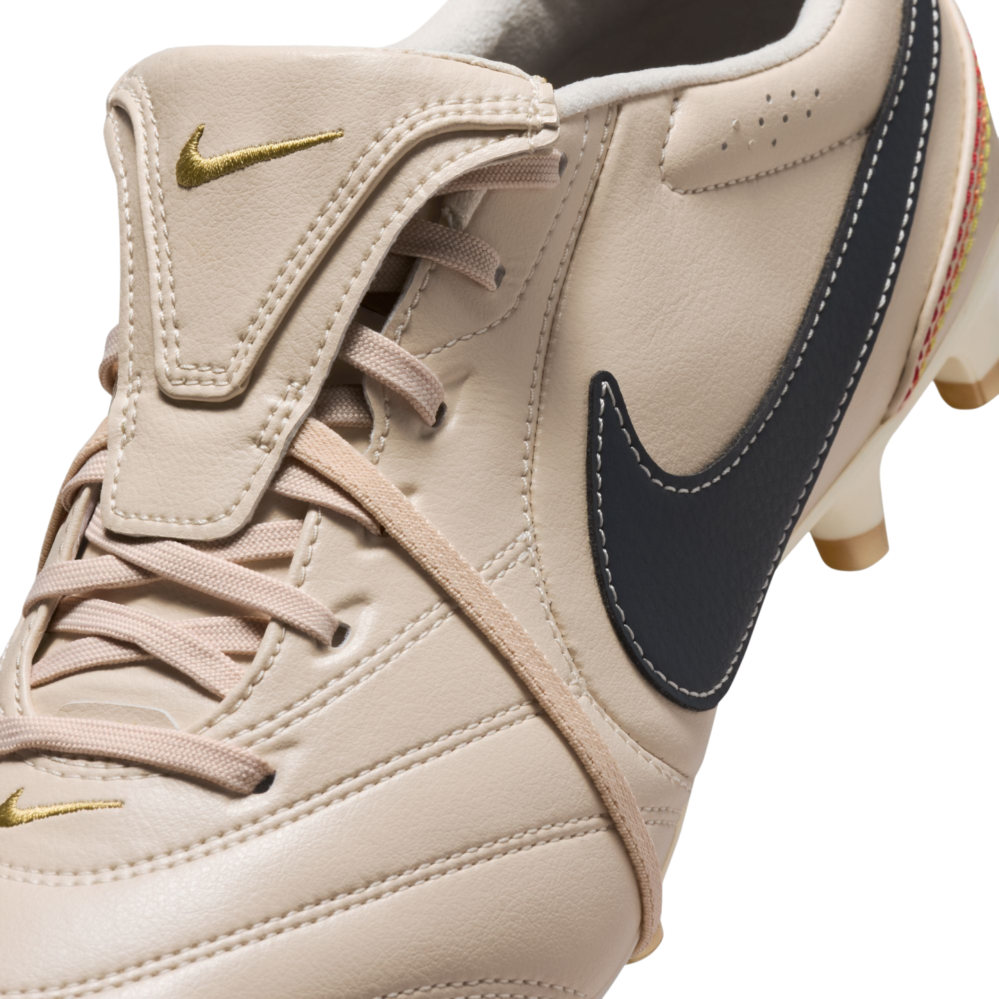 A close-up of a beige Nike soccer cleat featuring a fold-over tongue with a gold logo and a large navy blue side Swoosh with white stitching.