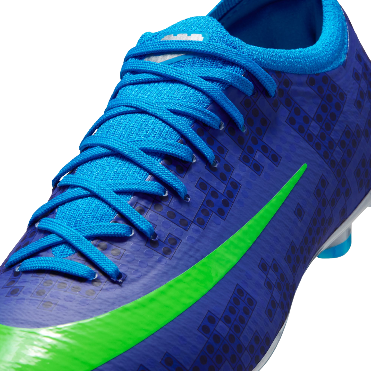 Close-up of a blue Nike soccer cleat with light blue laces, a bright green swoosh, and a dark blue LEGO brick pattern on the upper.