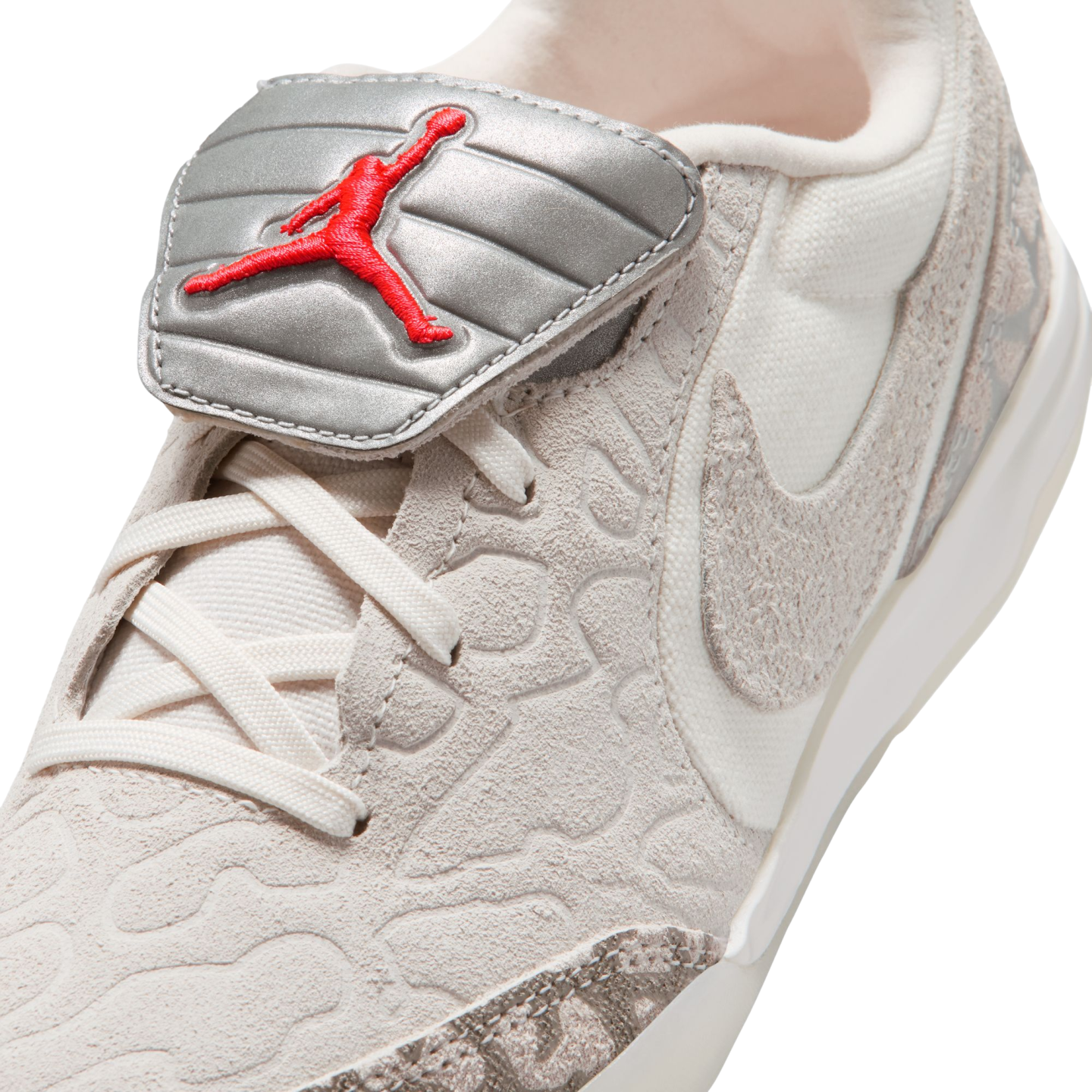 Close-up of a light beige sneaker featuring a metallic silver lace shroud with a red embroidered Jumpman logo. The shoe has a textured, patterned upper.