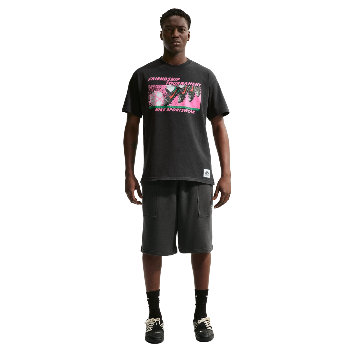 A man stands against a white background wearing a black Nike "Friendship Tournament" graphic t-shirt, charcoal shorts, and black sneakers.