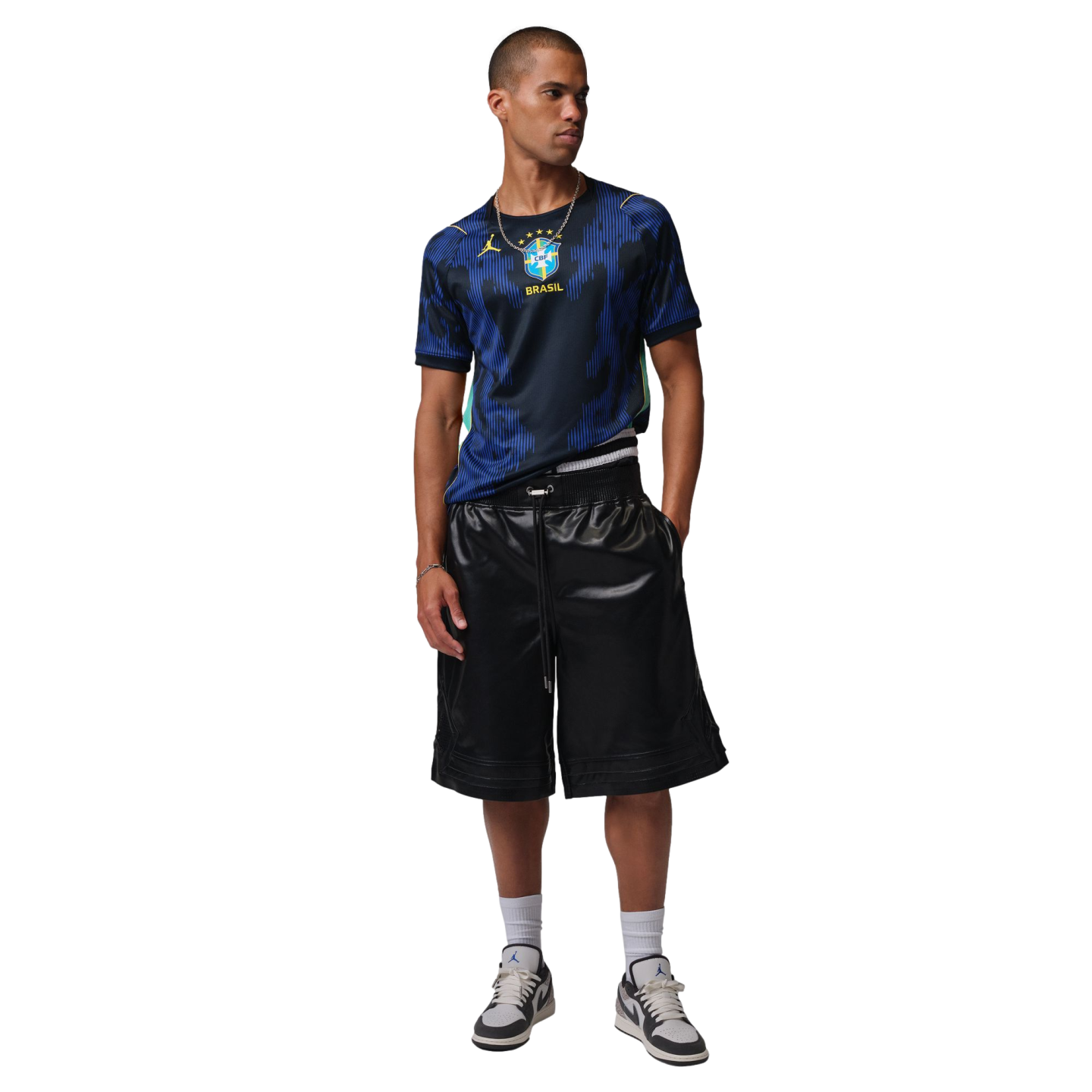 A male model wears a blue Brazil national team soccer jersey, black shiny shorts, and Jordan sneakers, standing against a black background.