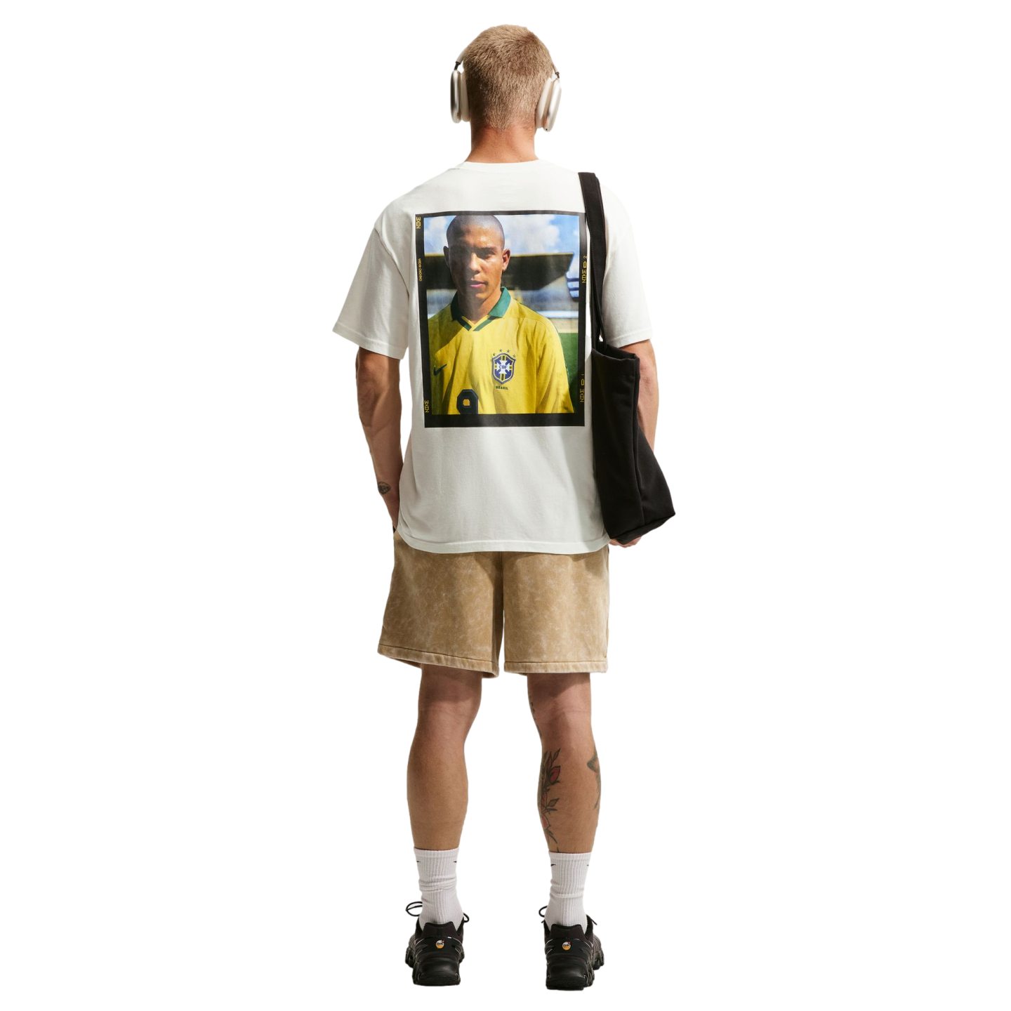 A rear view of a man with short blonde hair standing against a solid black background. He wears a white t-shirt with a large graphic print of a soccer player in a yellow Brazil jersey on the back, paired with tan shorts, white crew socks, and black sneake