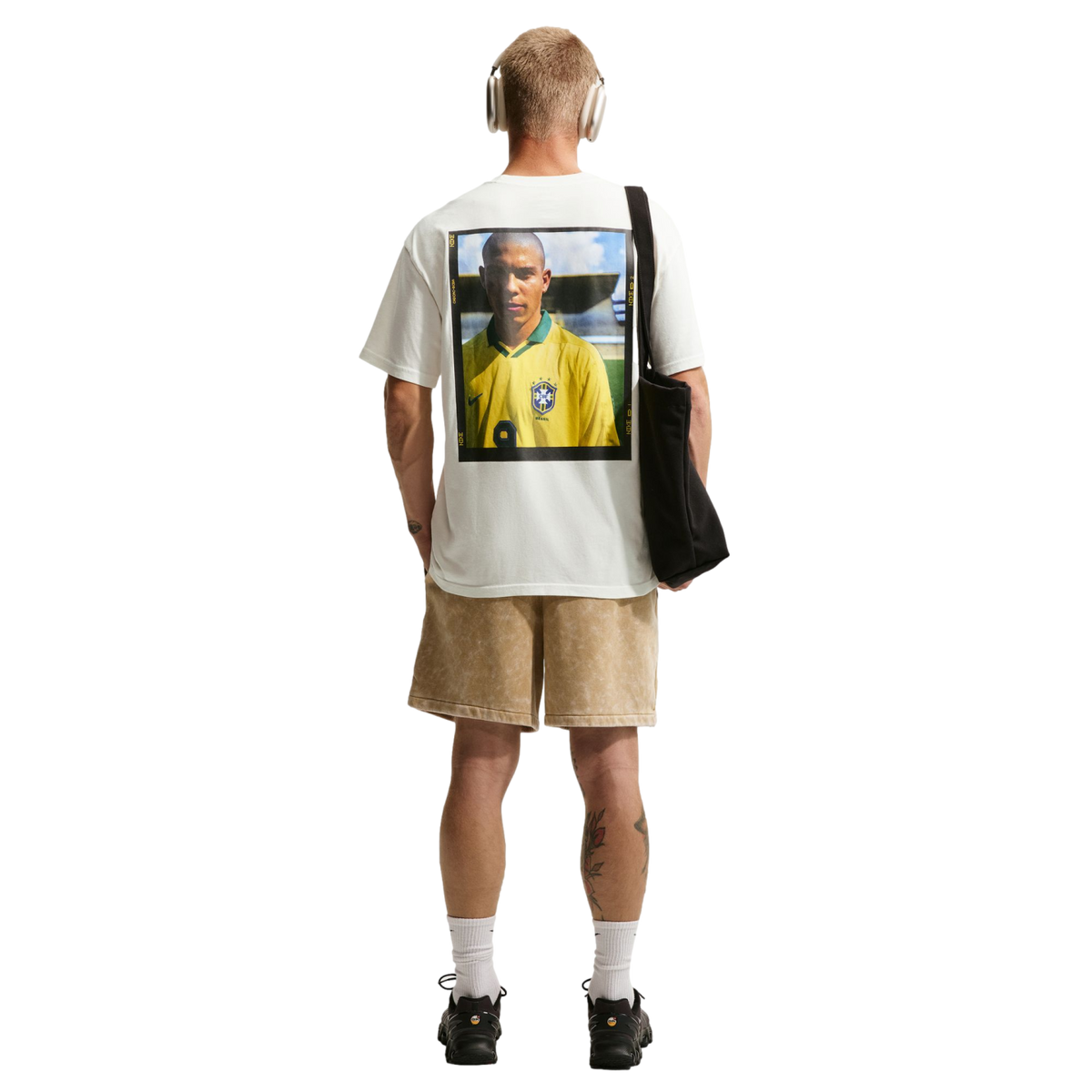 A rear view of a man with short blonde hair standing against a solid black background. He wears a white t-shirt with a large graphic print of a soccer player in a yellow Brazil jersey on the back, paired with tan shorts, white crew socks, and black sneake
