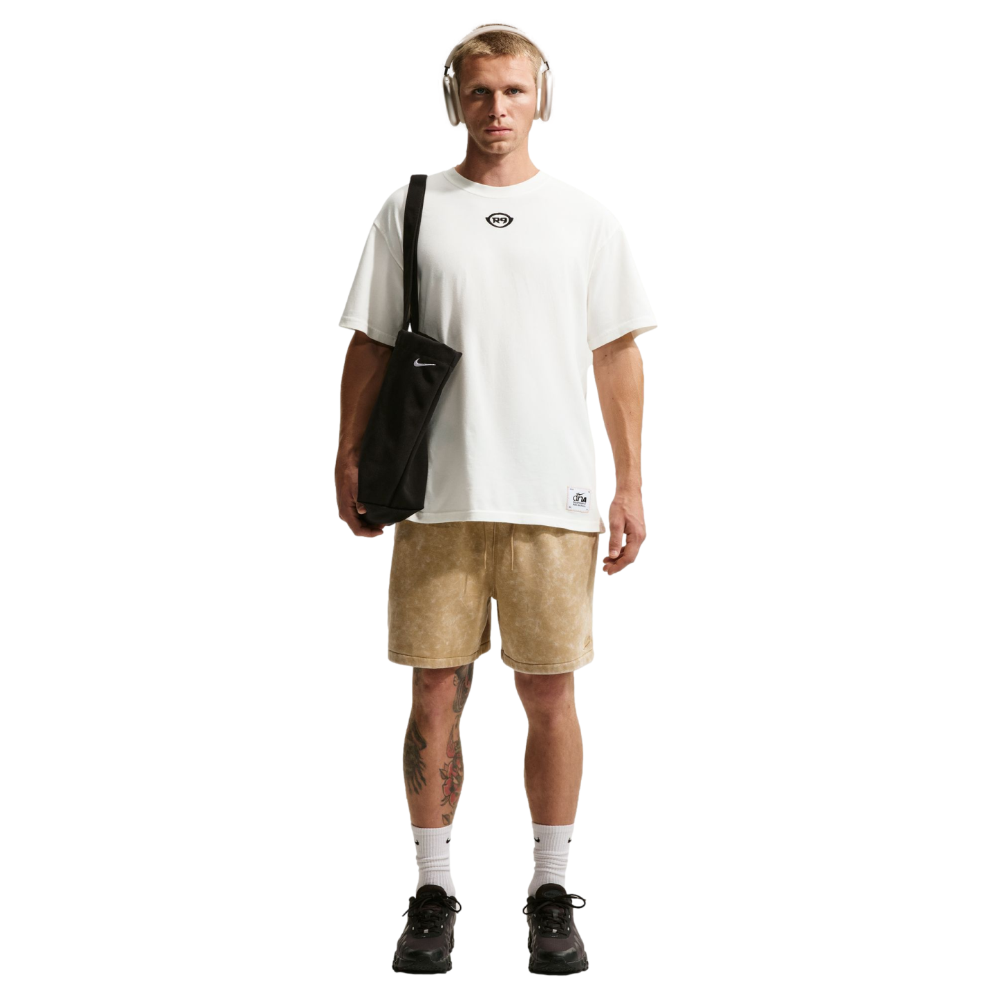 A man with short blonde hair and tan headphones stands against a black background, wearing an oversized white t-shirt and light brown patterned shorts. He carries a black Nike tote bag over his shoulder and wears white socks with black sneakers. Tattoos a