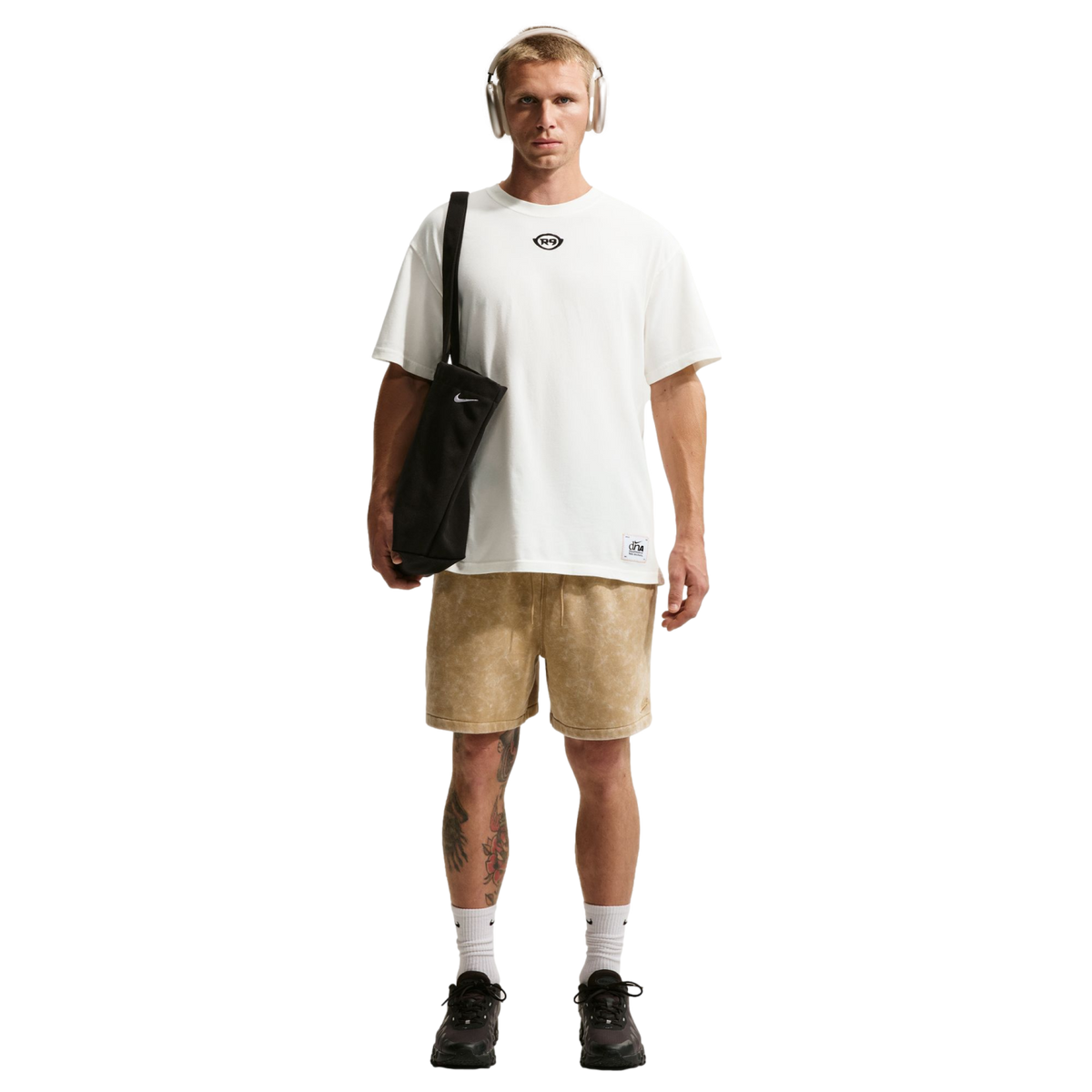 A man with short blonde hair and tan headphones stands against a black background, wearing an oversized white t-shirt and light brown patterned shorts. He carries a black Nike tote bag over his shoulder and wears white socks with black sneakers. Tattoos a
