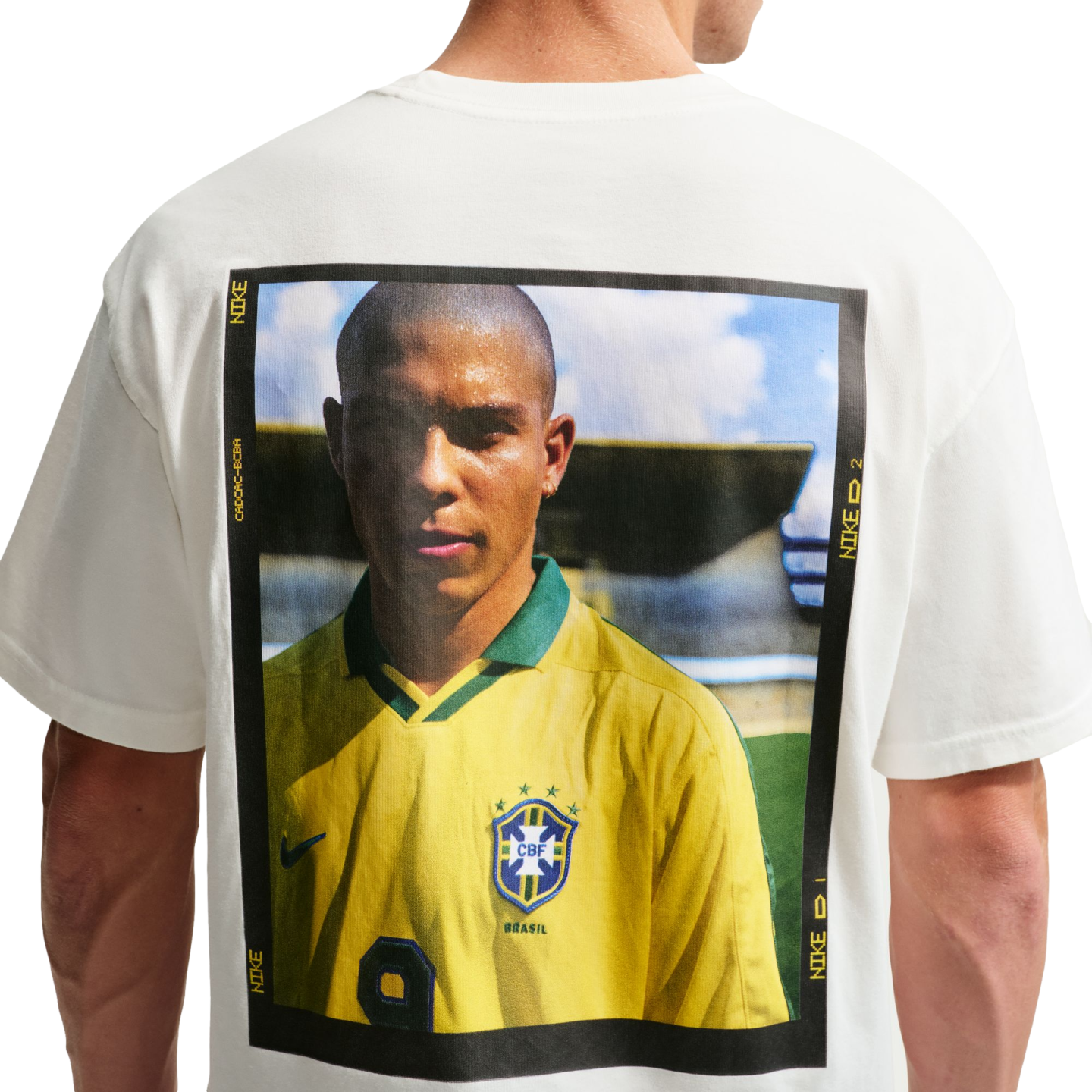 The back of a white t-shirt featuring a large, rectangular graphic print of Brazilian soccer legend Ronaldo Nazário. He is wearing a yellow Brazil national team jersey, and the portrait is enclosed in a black frame with yellow Nike branding text.
