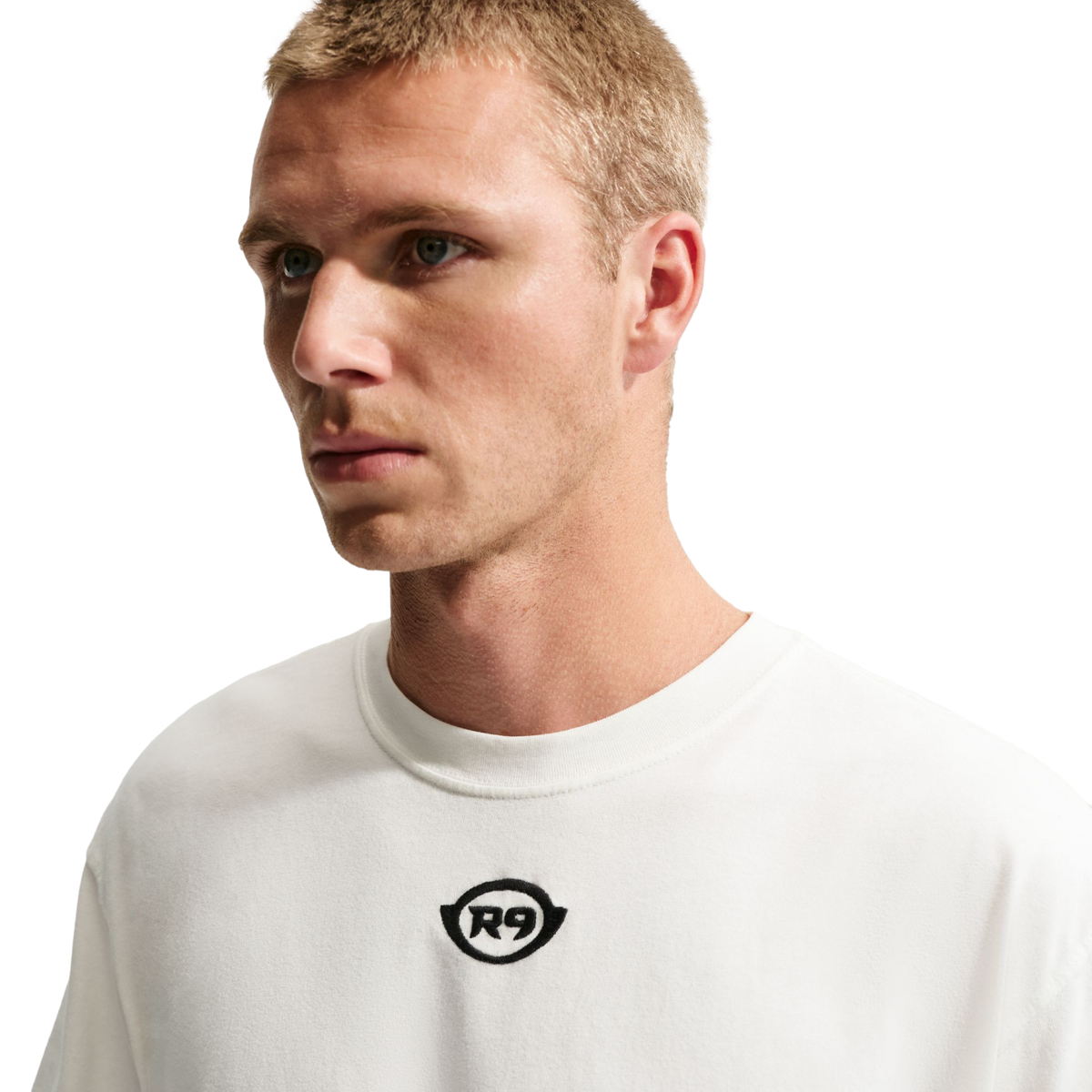 A medium shot of a man with short blonde hair wearing a white t-shirt with a small black "R9" logo in the center, set against a solid black background.