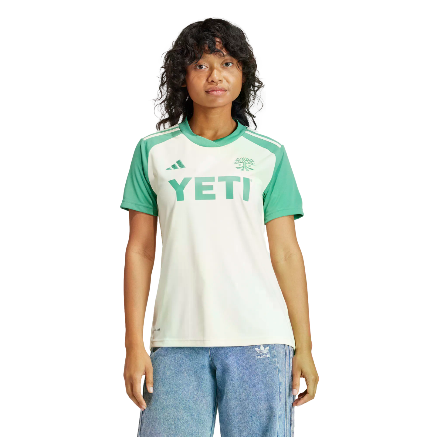 A person wears a white and green soccer jersey with a "YETI" logo and blue jeans against a black background.