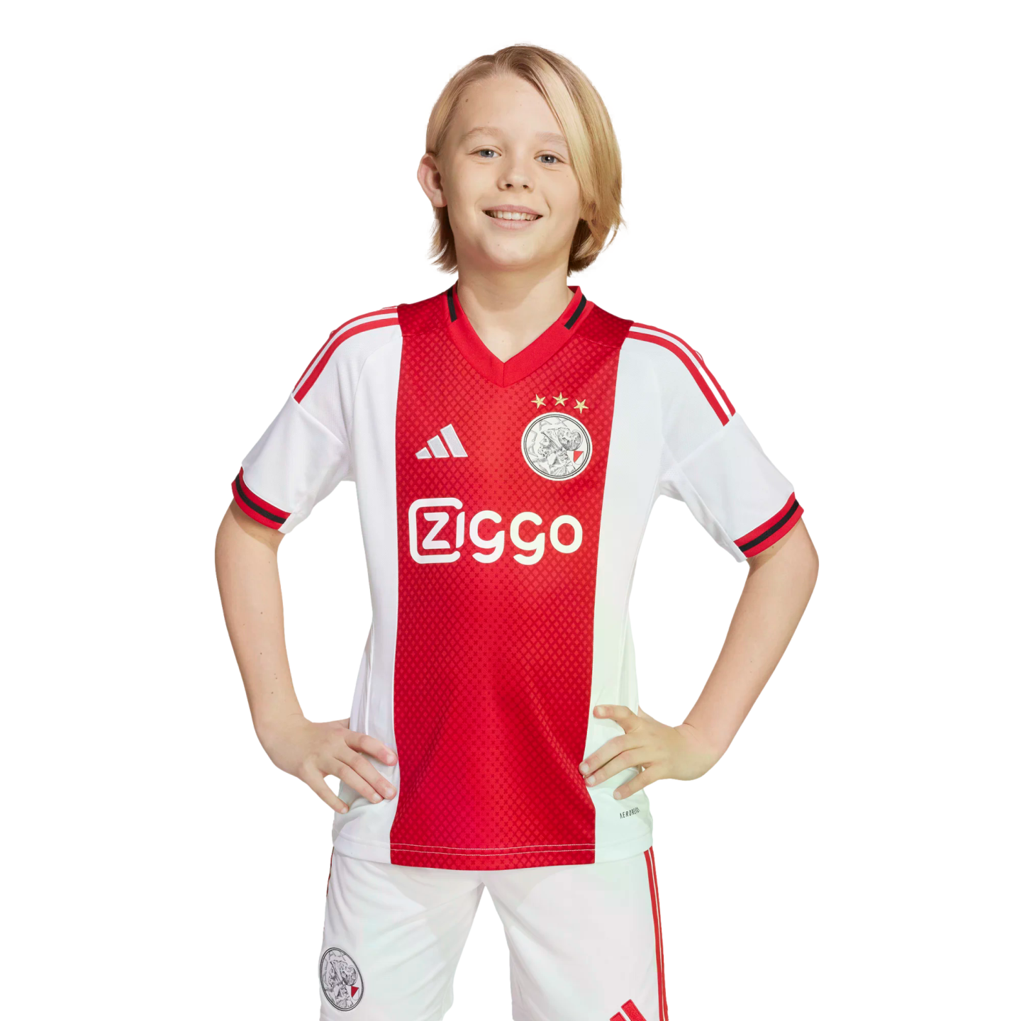 A smiling blond boy in a red and white Ajax home football kit stands with hands on his hips against a plain white background.