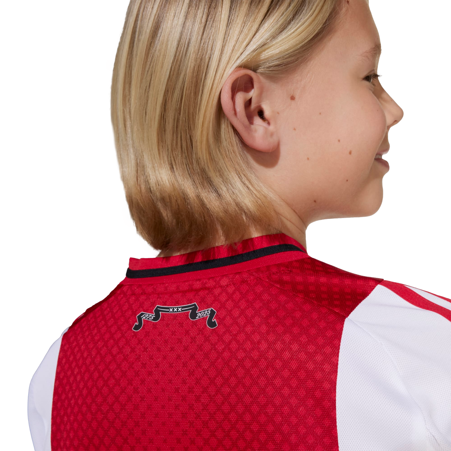 A blonde child from behind wearing a red and white soccer jersey. A small black banner on the collar reads "1275 XXX 2025".