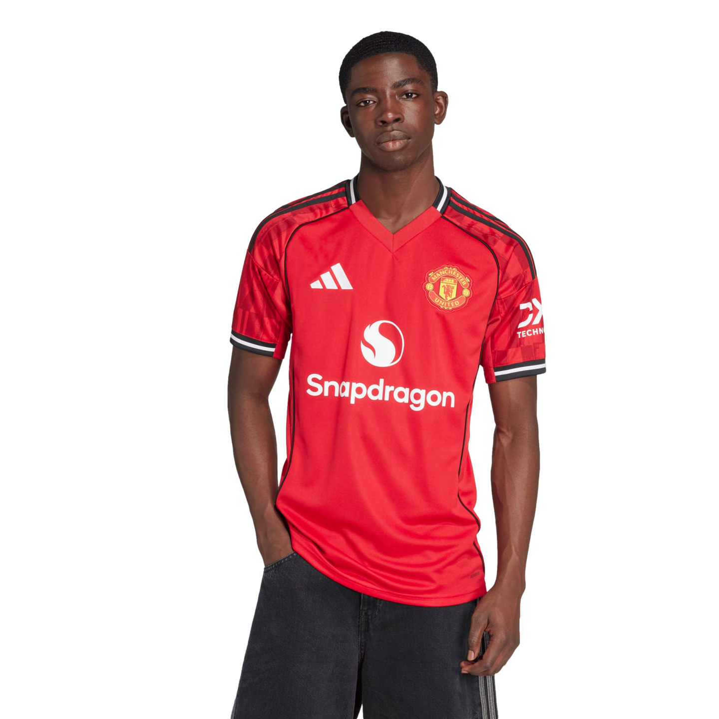 A Black man stands against a white background wearing a red Manchester United home jersey with the Snapdragon logo and black pants.