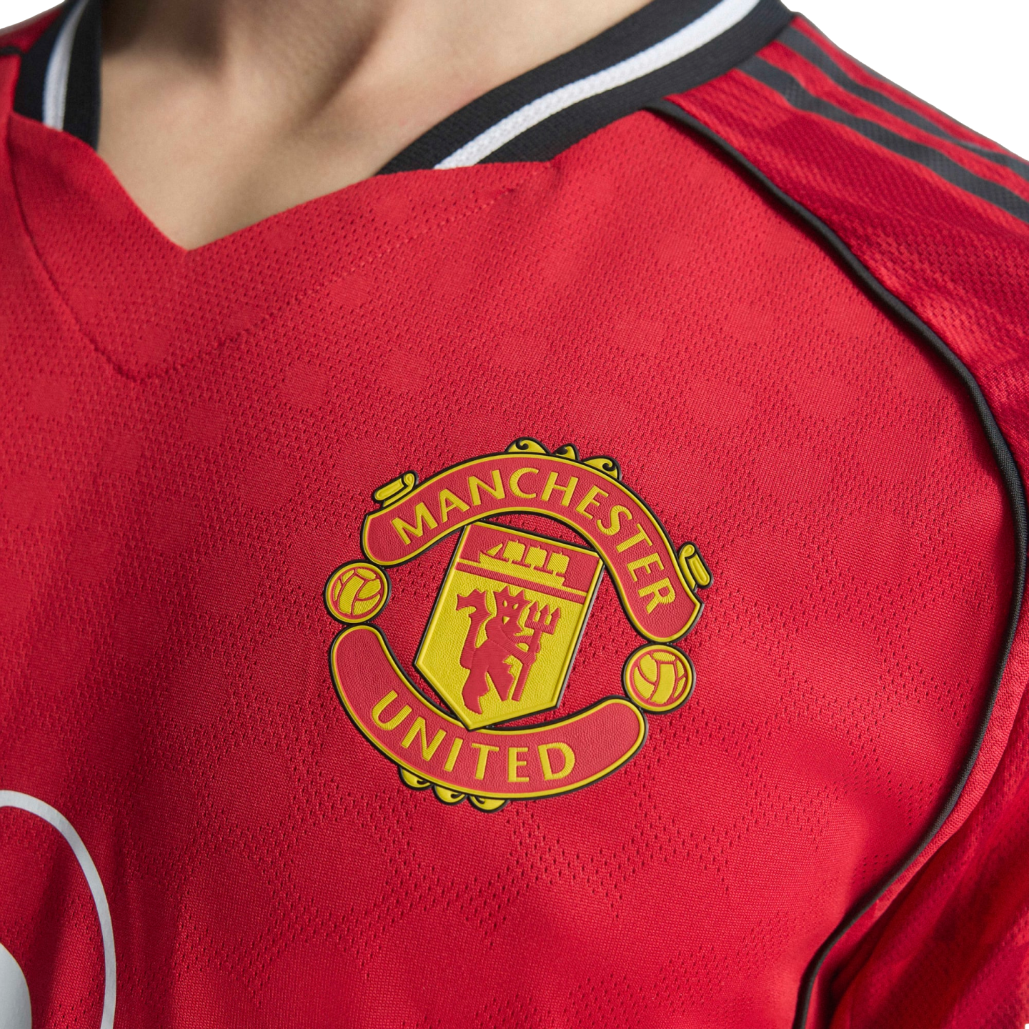 Close-up of a red Manchester United football jersey featuring the team's yellow and red crest on the chest, with black shoulder stripes and patterned fabric.
