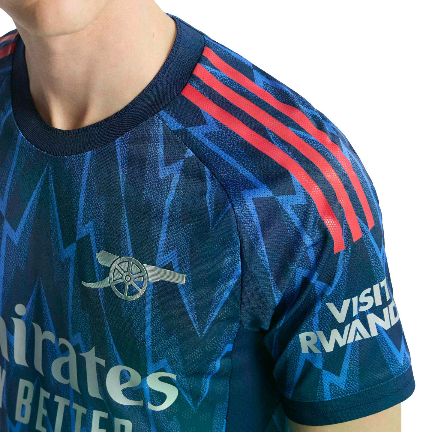 Close-up of a blue Arsenal soccer jersey with a zig-zag pattern, red shoulder stripes, a silver cannon crest, and "Visit Rwanda" sleeve logo.