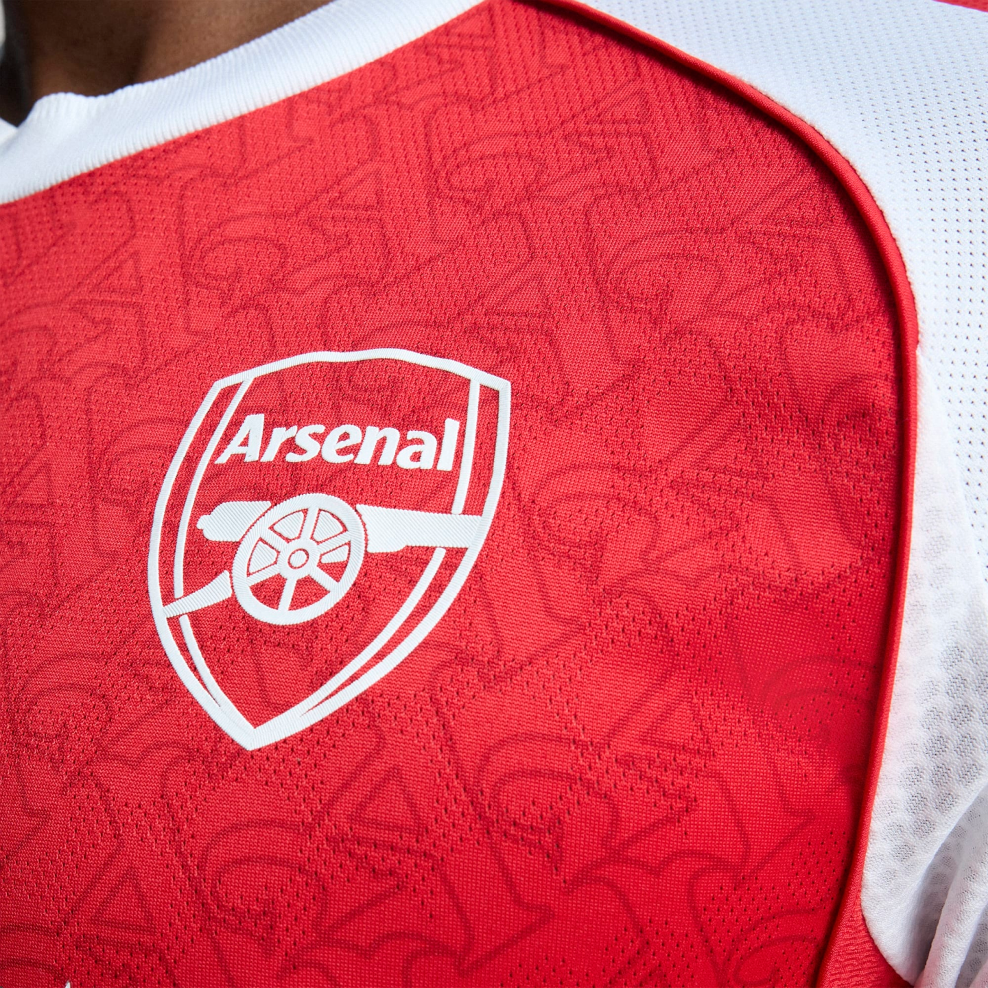 Close-up of a red Arsenal home jersey featuring the white club crest on a textured fabric with a subtle repeating pattern and white shoulder trim.