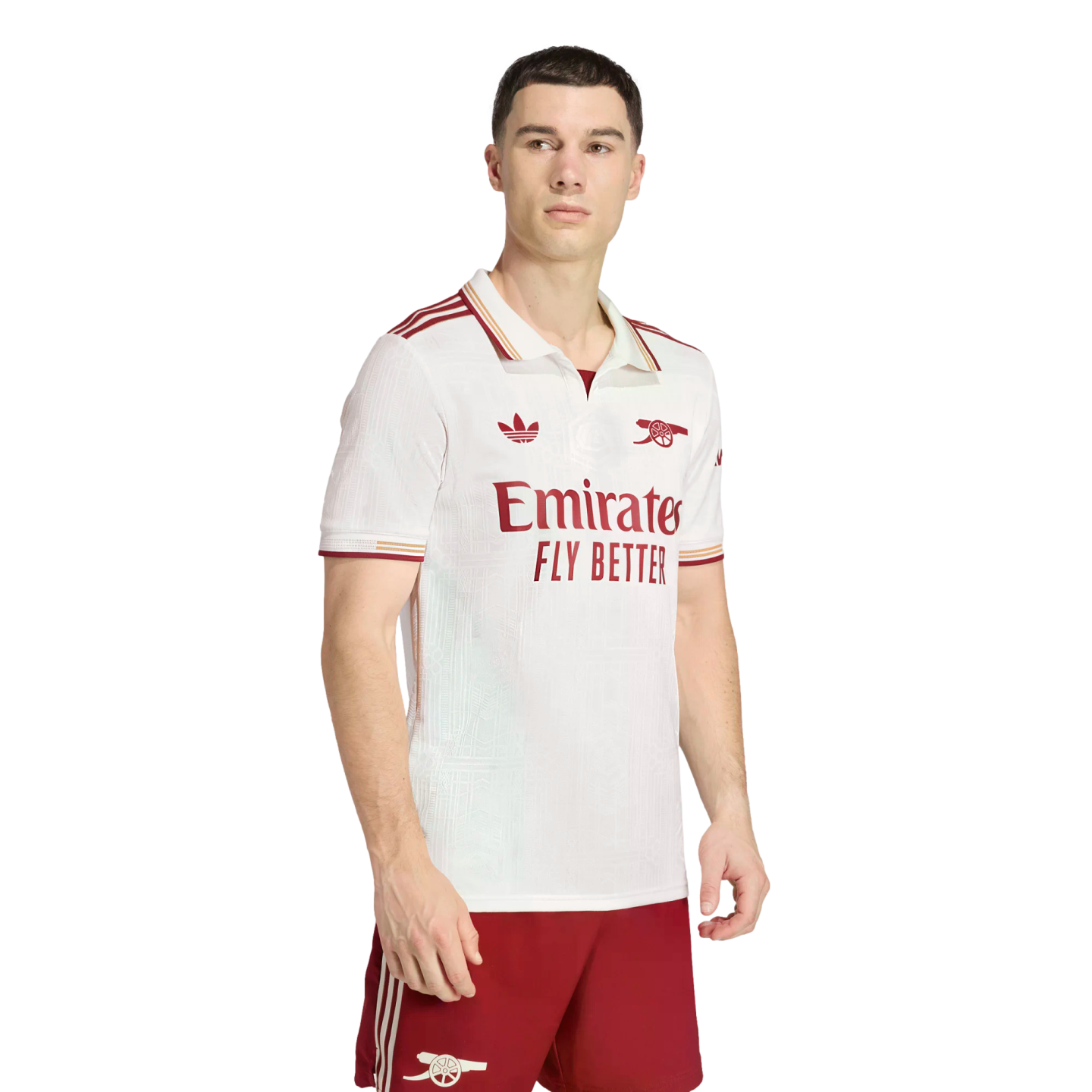 A man models the Arsenal 2024/25 third kit, featuring a white collared jersey with maroon accents and the Adidas trefoil logo, paired with maroon shorts against a black background.