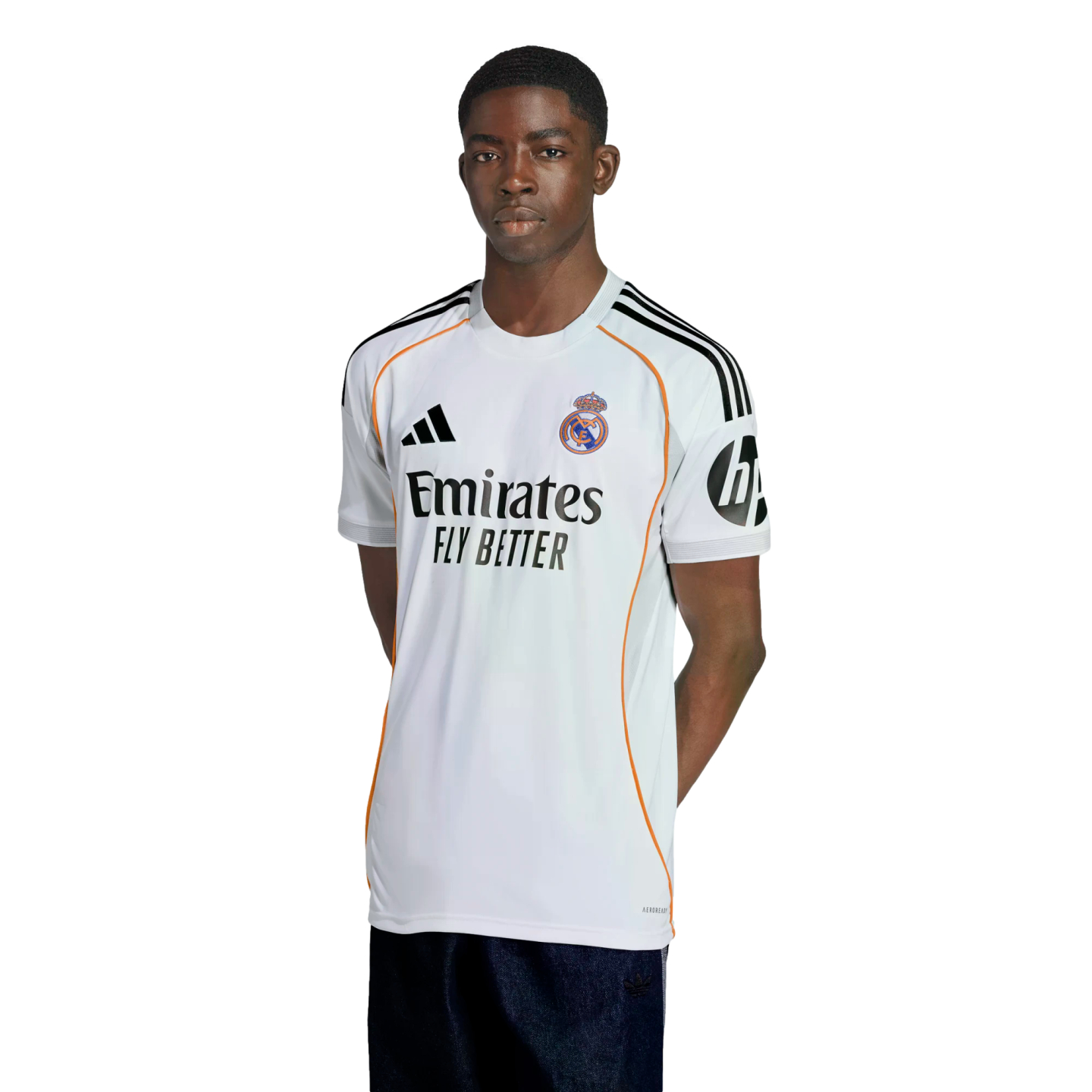 A young Black man in a white Real Madrid soccer jersey with orange accents and sponsor logos, standing with hands behind his back against a white background.